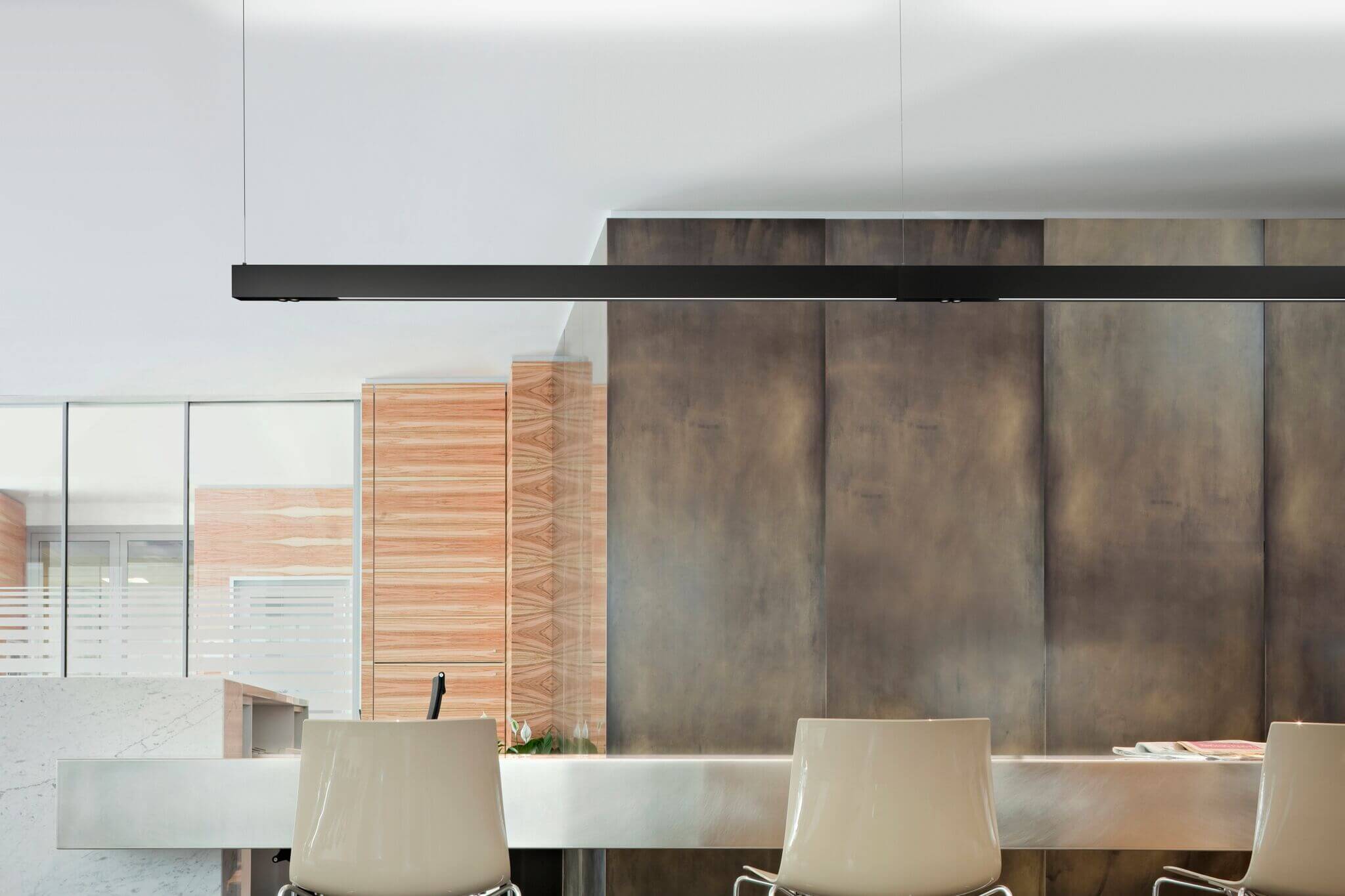 Minimalistischer Innenraum mit langer, schwarzer Pendelleuchte über einer hellen Theke. Im Vordergrund stehen zwei weisse Barstühle. Im Hintergrund eine grossflächige Wandverkleidung in Betonoptik sowie Glas und Holz Elemente, die den Raum klar strukturieren.