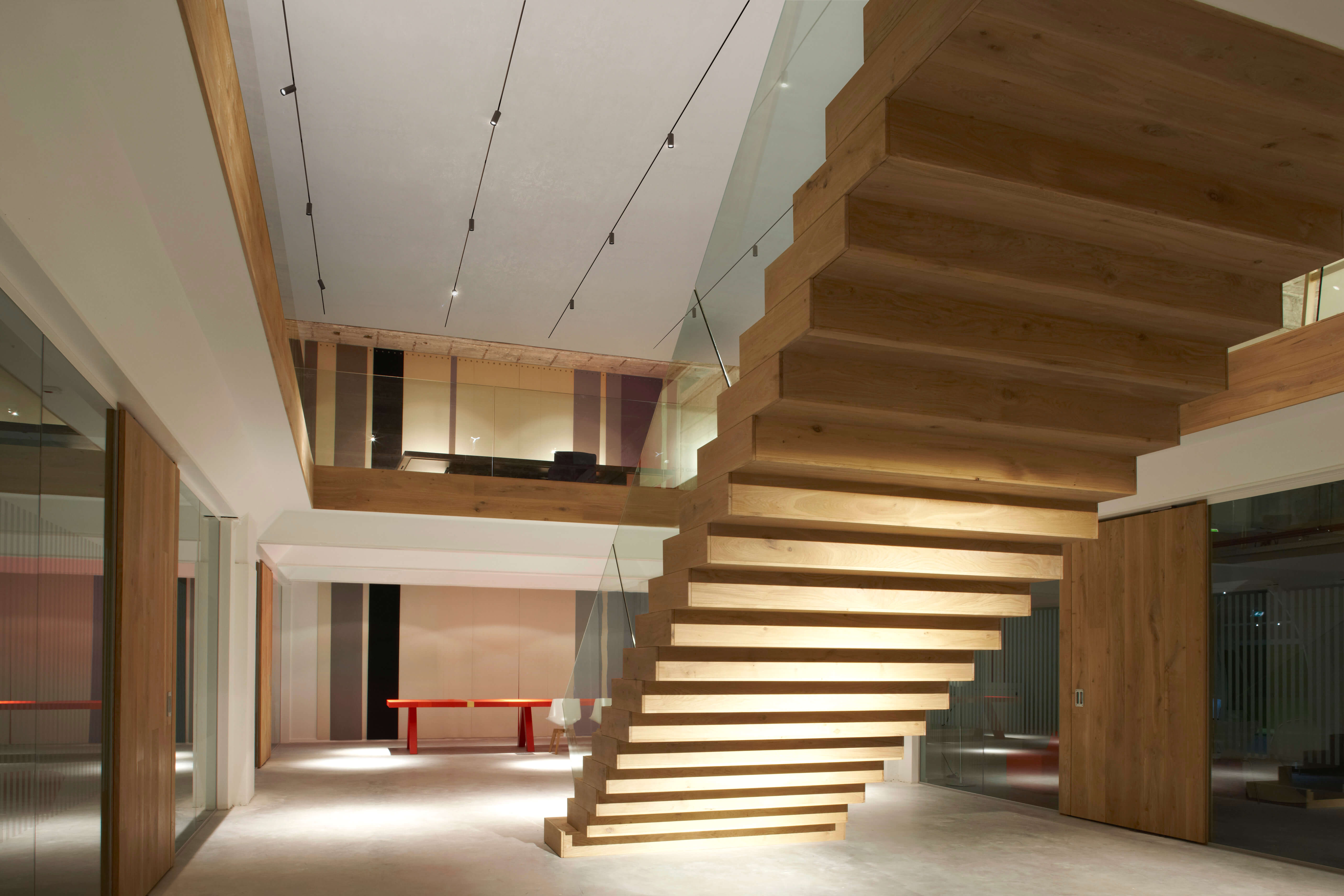 Moderne Innenarchitektur mit skulpturaler Holztreppe und integrierter Stufenbeleuchtung. Glasgeländer und offene Galerieebene betonen die Raumhöhe. Klare Wände, grosse Holzflächen und dezente Decken Spots schaffen eine ruhige, hochwertige Atmosphäre. Im Hintergrund steht ein roter Tisch als Farbakzent.