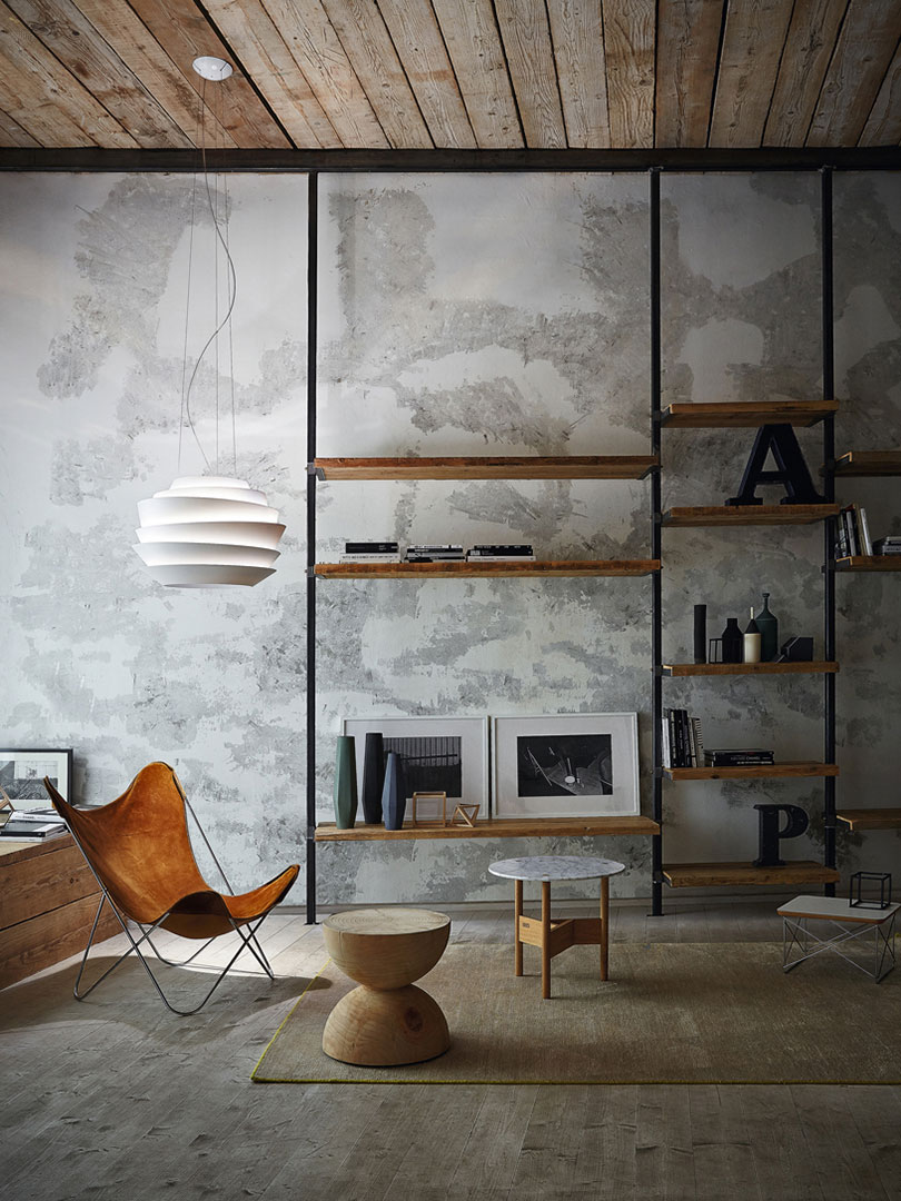 Wohnbereich mit hoher Regalwand aus schwarzem Metall und Holzablagen vor einer rauen Betonoptik Wand. Links hängt eine weisse Pendelleuchte über einem Lounge Sessel in Cognac Leder. Auf den Regalen stehen Bücher, Vasen und gerahmte Bilder, rechts sind dekorative Buchstaben platziert. Holzbalkendecke und Teppich ergänzen das industrielle Setting.