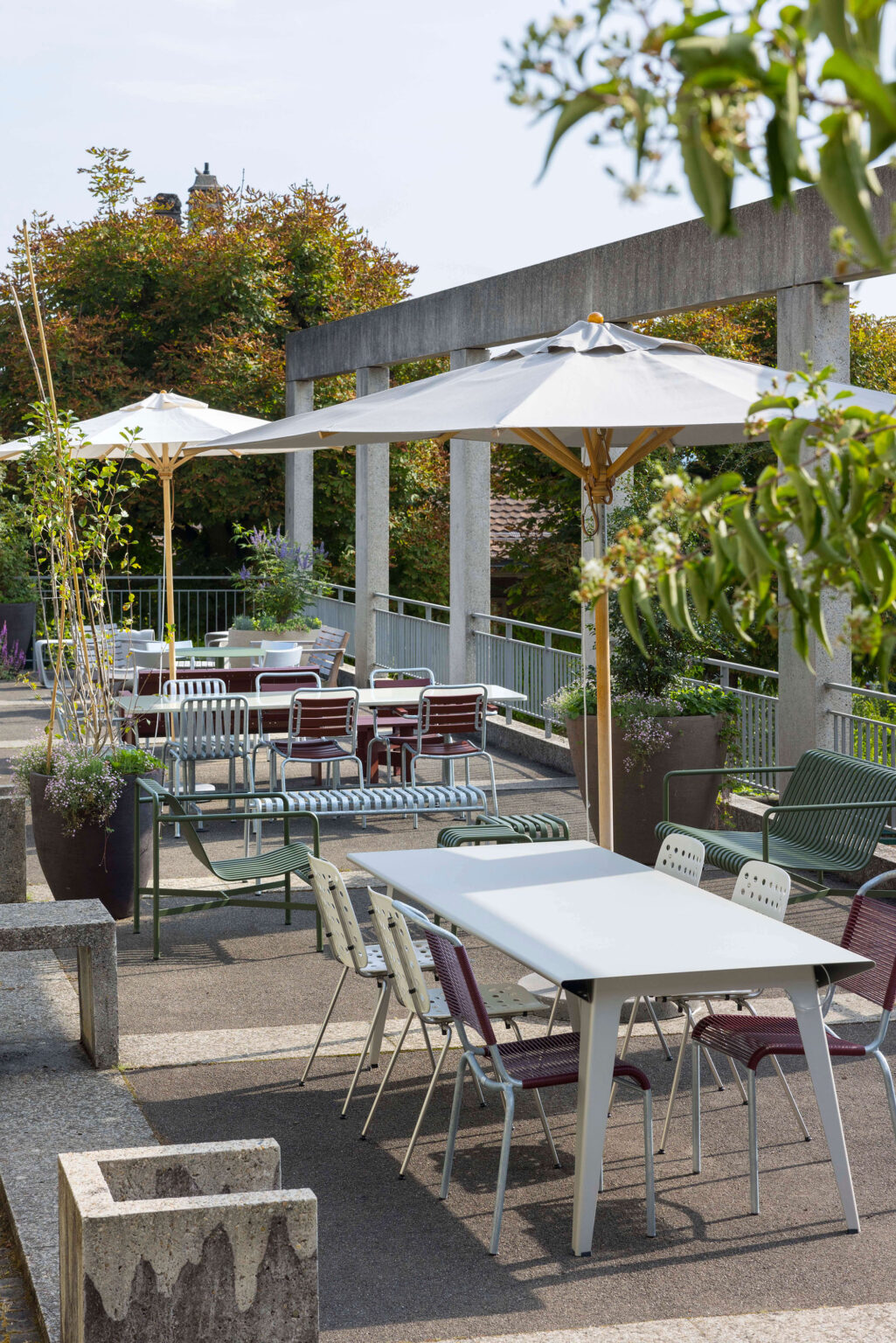 Terrasse mit weissen Tischen, gemischten Outdoor-Stühlen und Bänken, grossen Sonnenschirmen und Pflanzkübeln entlang einer Betonkolonnade auf dem Areal der Friedenskirche in Bern.