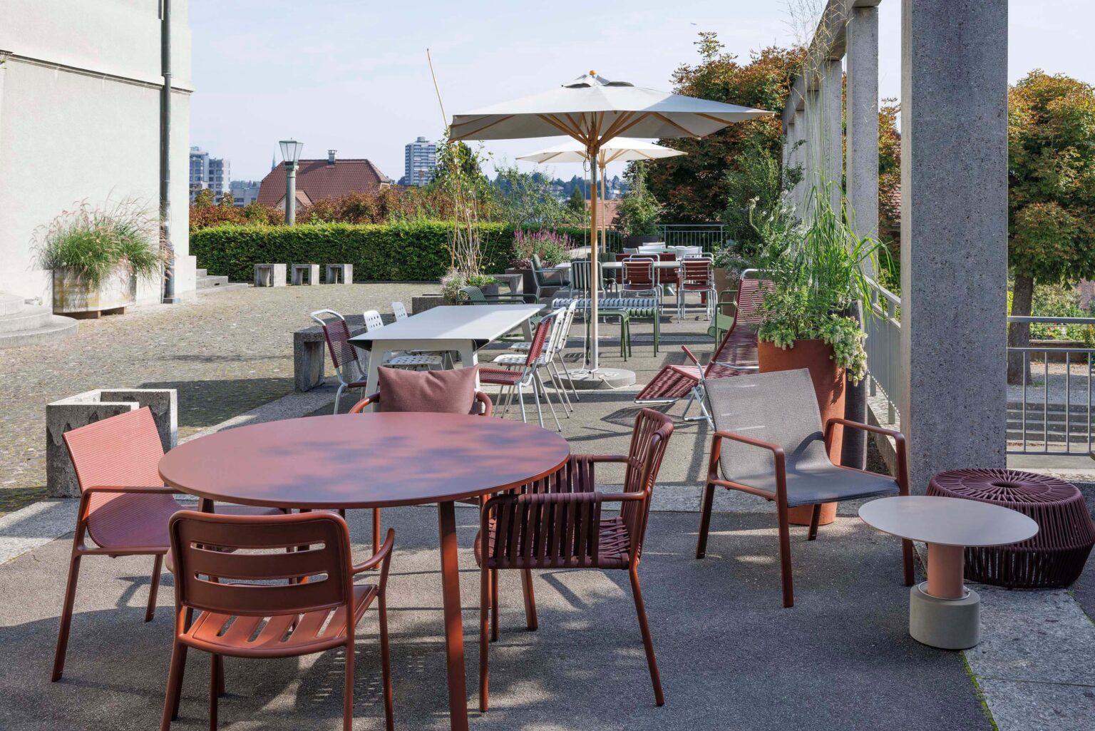 Grosszügiger Aussenbereich mit runden und rechteckigen Tischen, Lounge-Sesseln und Sonnenschirmen neben einer Betonarkade auf dem Areal der Friedenskirche in Bern.