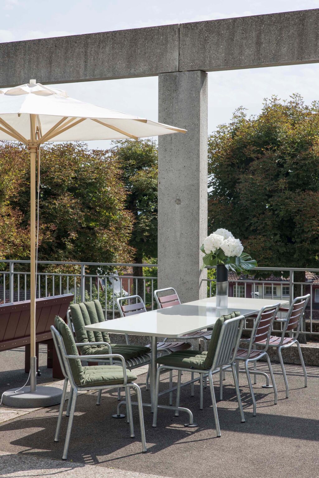 Outdoortisch mit Metallstühlen und grünen Sitzkissen, Hortensienvase als Mittelpunkt unter Sonnenschirm an der Betonkolonnade auf dem Areal der Friedenskirche in Bern.