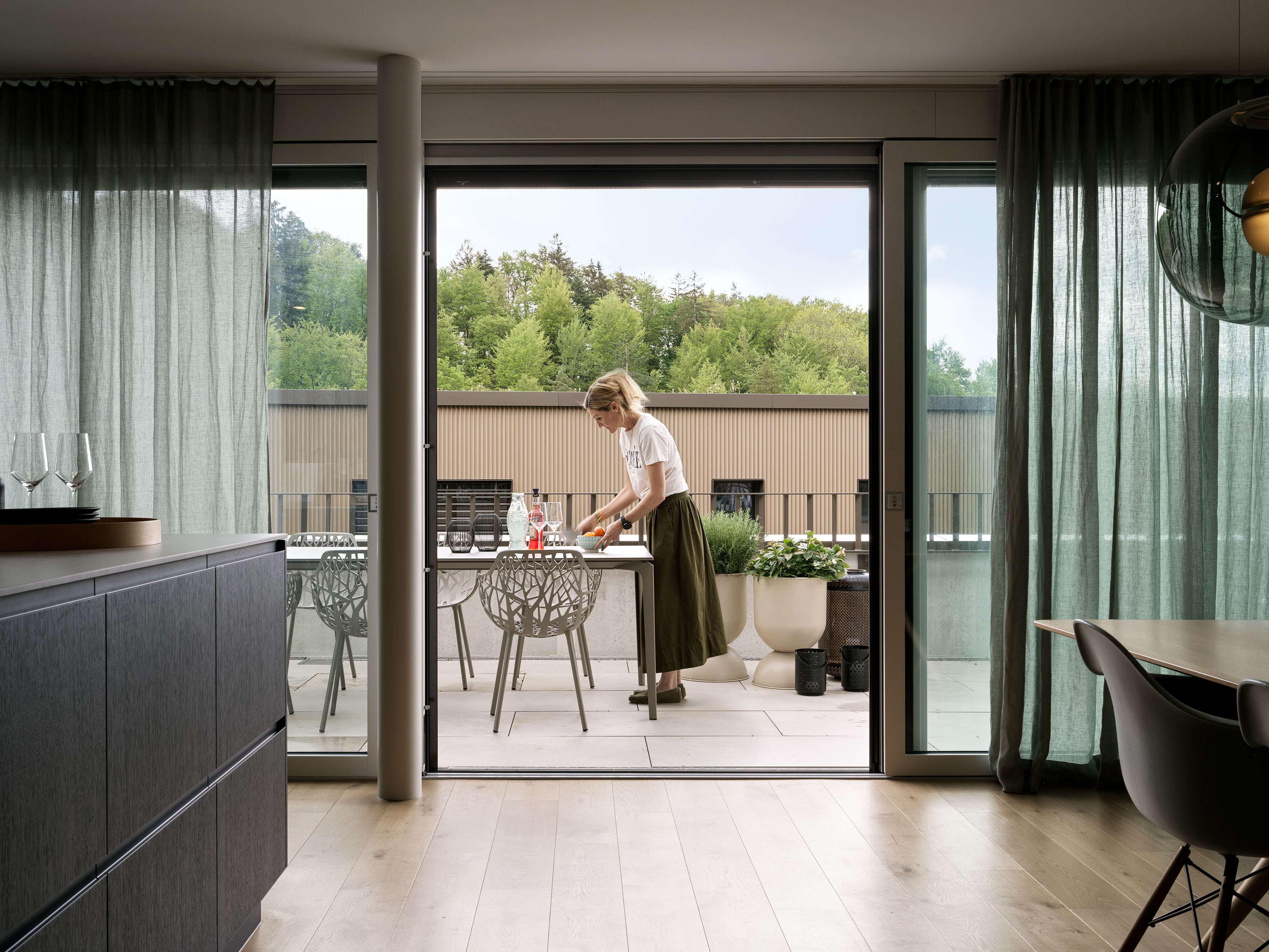 Blick vom Wohn-/Essbereich auf die Terrasse: offene Schiebetüren mit grünen Vorhängen, heller Holzboden; innen dunkles Sideboard und Schalenstühle.
