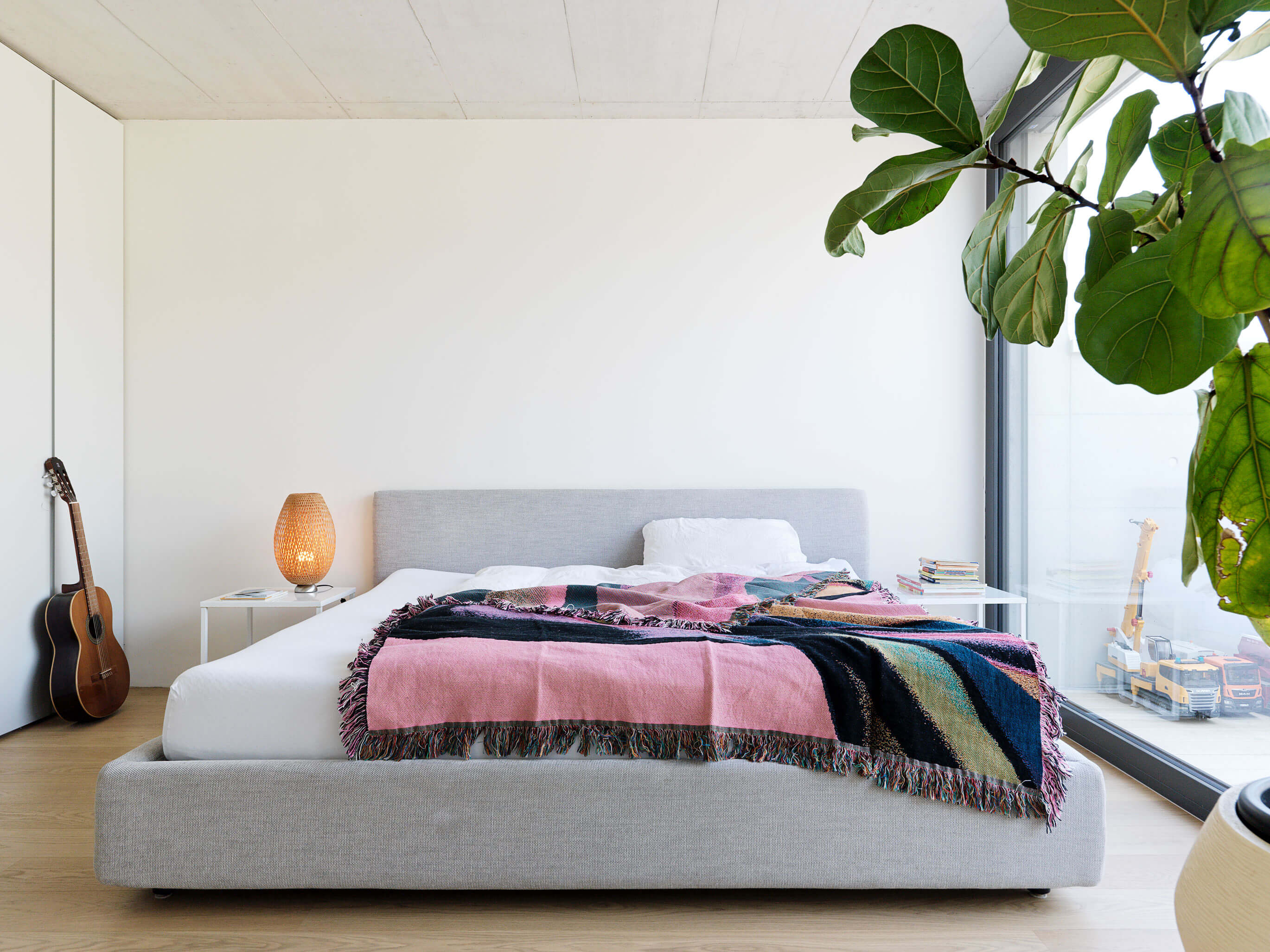 Helles Schlafzimmer mit grauem Polsterbett vor raumhoher Verglasung, farbiger Decke, Nachttischleuchte und grosser Zimmerpflanze, im Patiohaus in Winterthur.