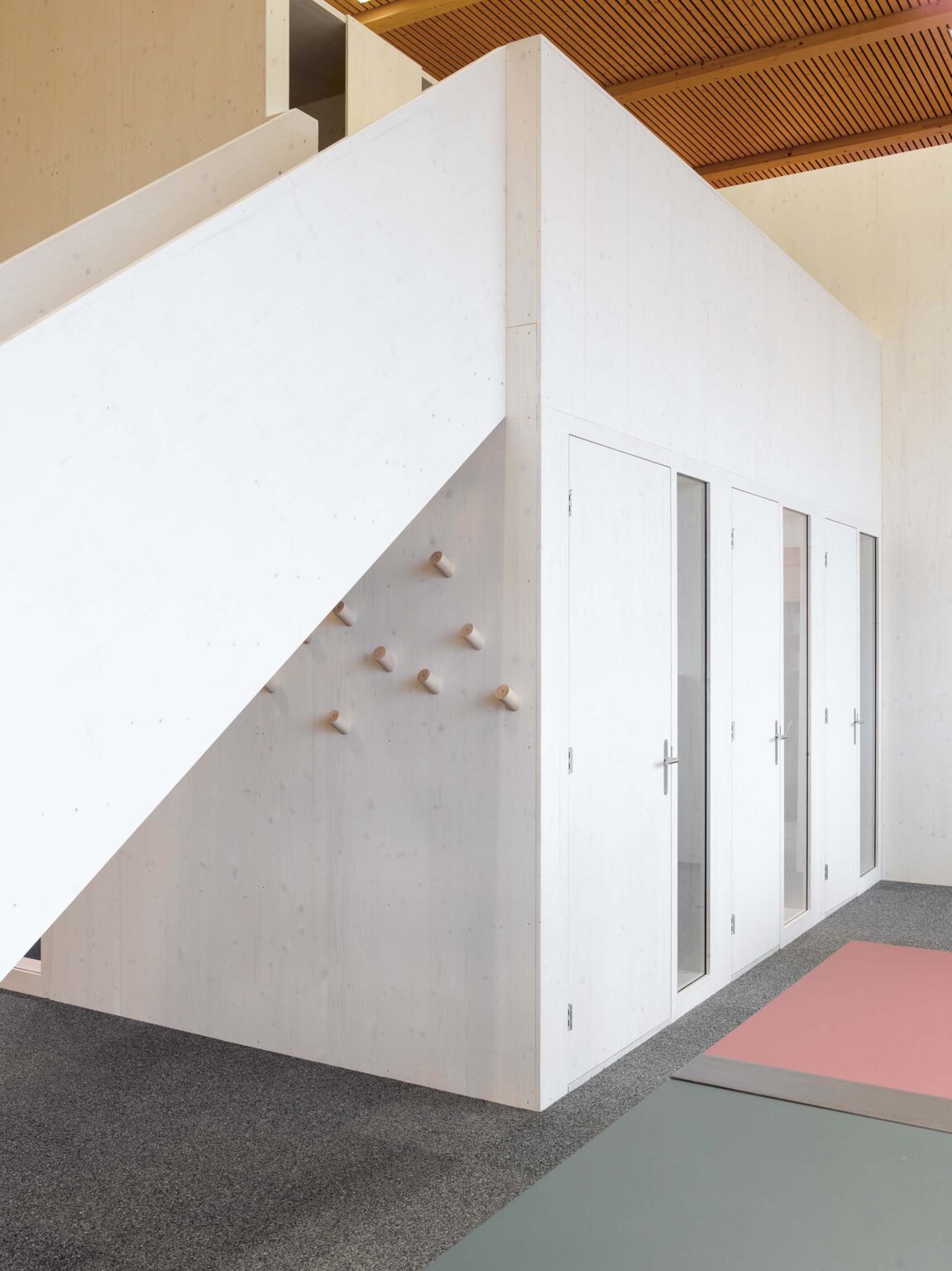 Innenraum der zur Lernhalle umgenutzten Turnhalle in Orpund mit weissem Holzvolumen, Treppe, Garderobenhaken und Türen, auf grauem Boden mit farbigen Matte.