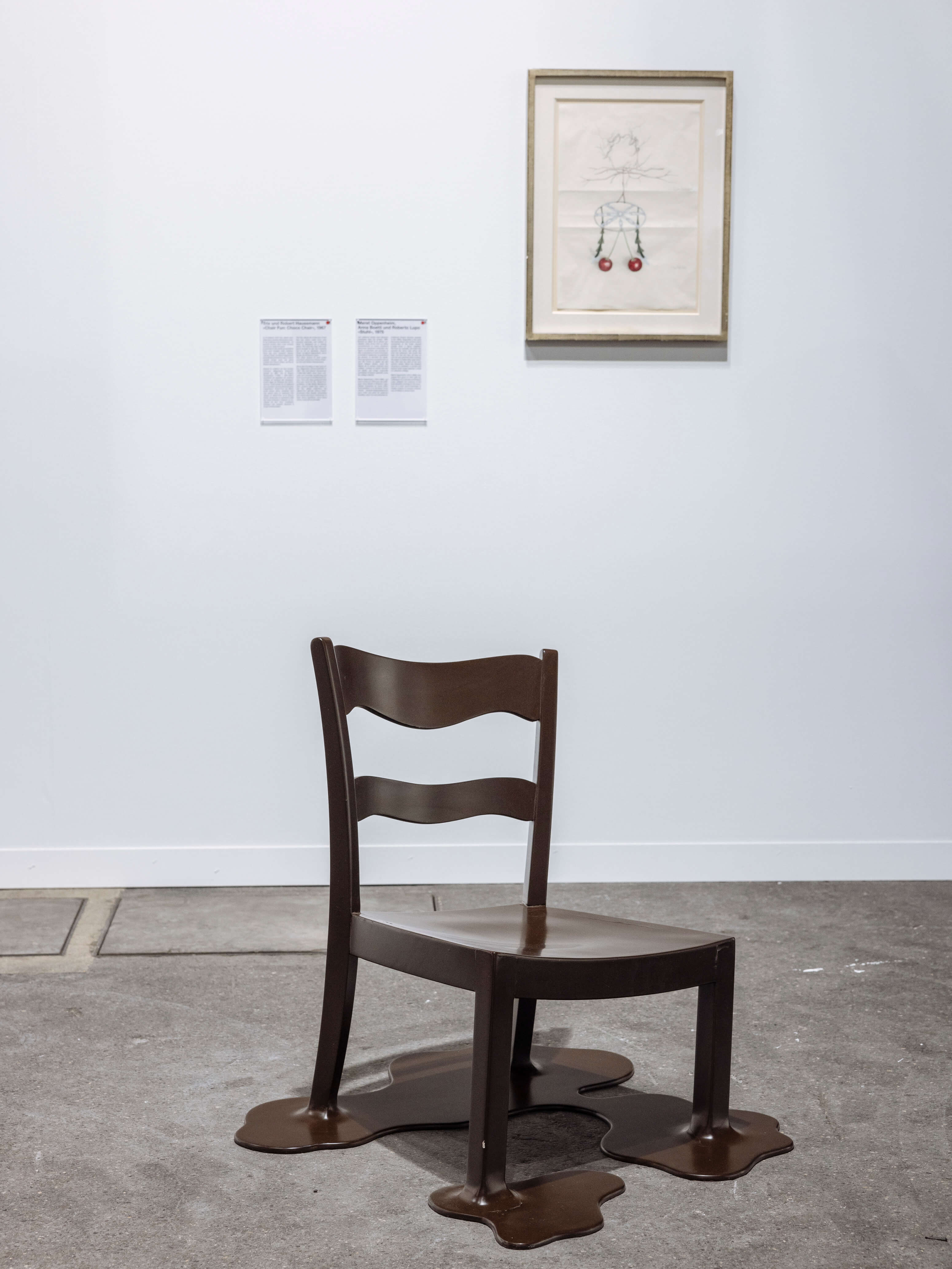 Ausstellungsansicht mit braunem Stuhl, dessen Füsse wie schmelzend zu Pfützen verlaufen. An der weissen Wand dahinter hängt eine gerahmte Zeichnung mit zwei roten Kirschen sowie zwei Wandtexte.