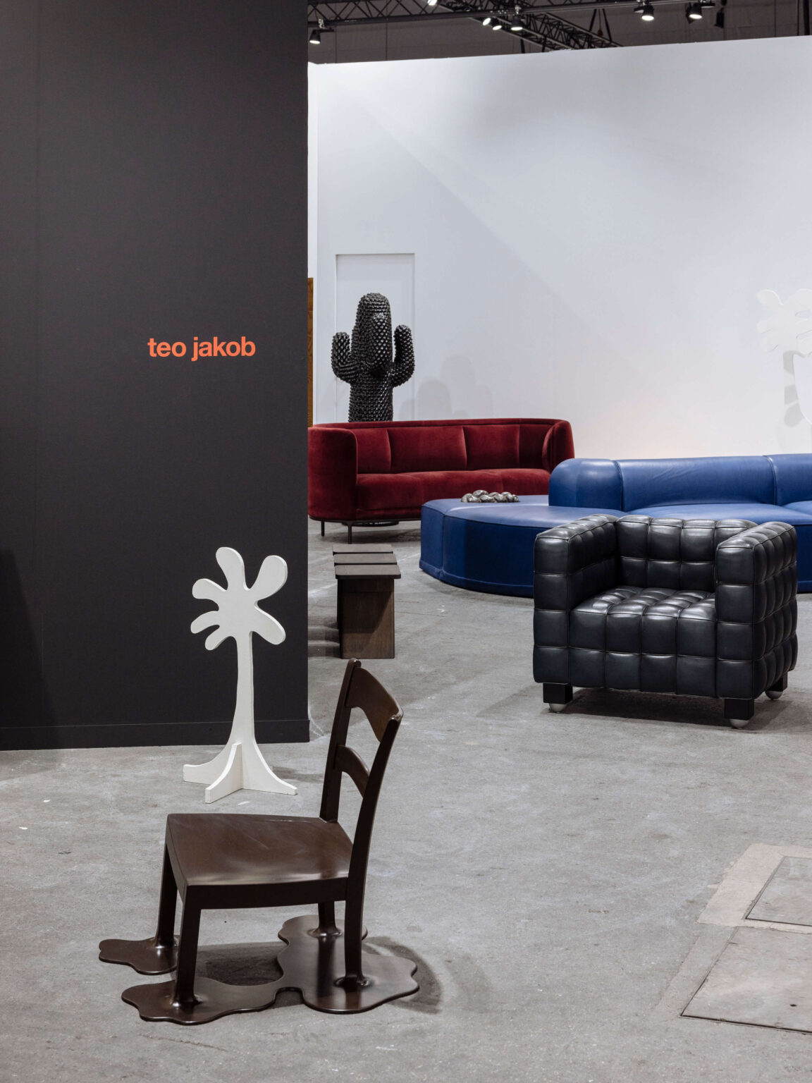 Messestand von teo jakob mit dunkler Wand und Logo links. Im Vordergrund ein brauner Stuhl mit „schmelzender“ Basis und eine weisse, palmenartige Garderobe. Dahinter rote Sofas, blaue Rundbank, schwarzer gesteppter Sessel und eine kaktusförmige Skulptur.