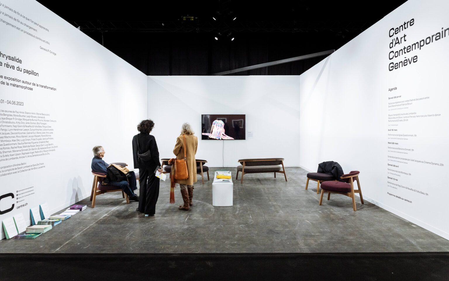 Ausstellungsraum des Centre d’Art Contemporain Genève mit weissen Wänden und Wandtexten, mittig ein Monitor mit Video, Sitzbänke und Stühle, zwei Besucherinnen stehen, eine Person sitzt und liest, Broschüren auf Podest und Boden.
