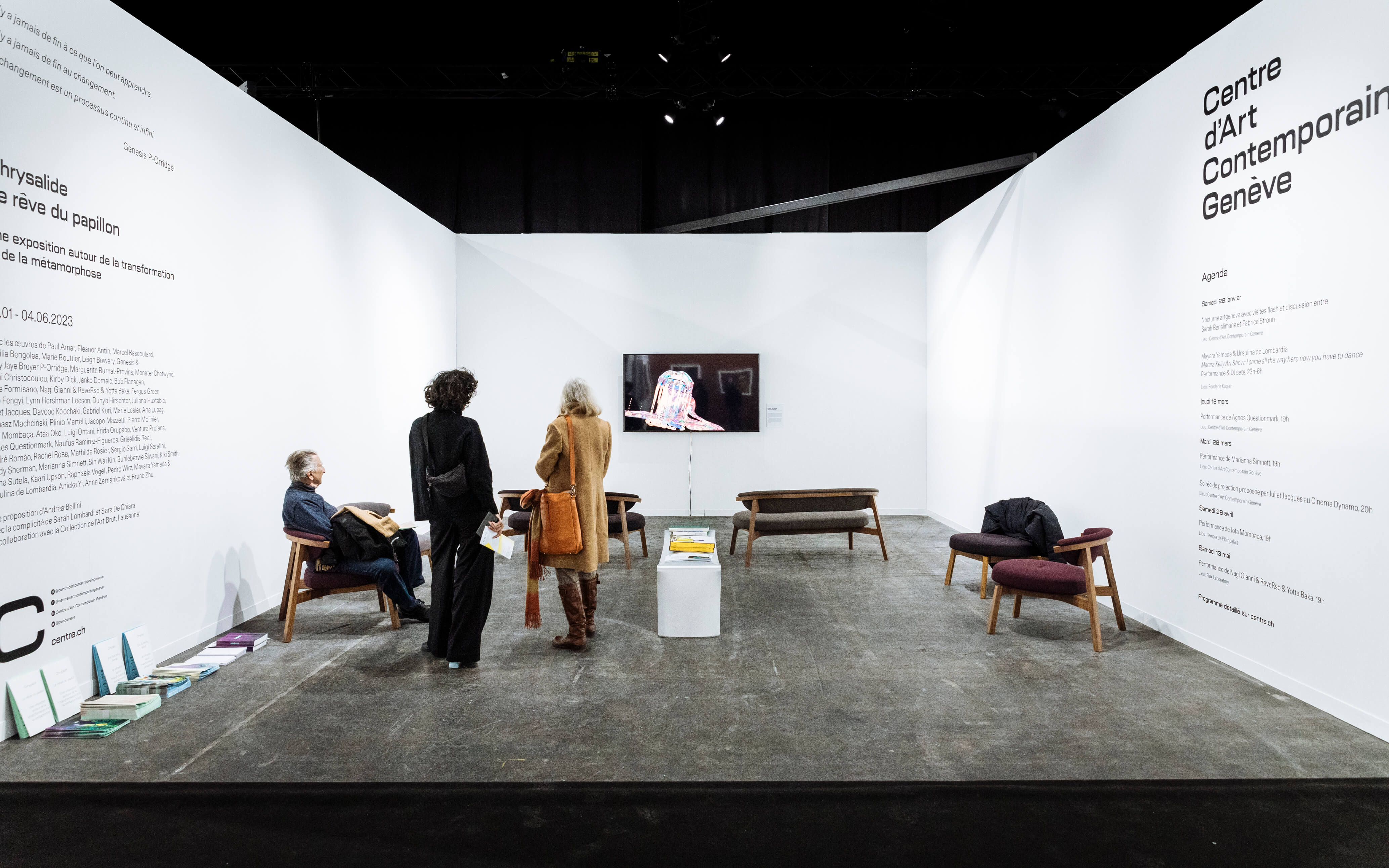 Ausstellungsraum des Centre d’Art Contemporain Genève mit weissen Wänden und Wandtexten, mittig ein Monitor mit Video, Sitzbänke und Stühle, zwei Besucherinnen stehen, eine Person sitzt und liest, Broschüren auf Podest und Boden.