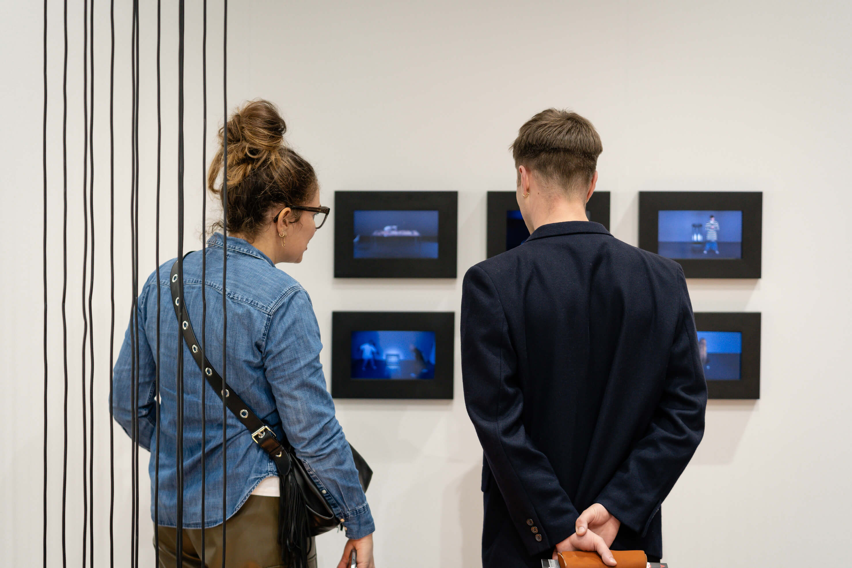 Zwei Personen betrachten in einer Ausstellung mehrere kleine, blau leuchtende Videomonitore an einer weissen Wand; links eine Person im Jeanshemd mit Umhängetasche, rechts eine Person im dunklen Blazer mit hinter dem Rücken verschränkten Händen, vorn verlaufen schwarze Installationsschnüre.
