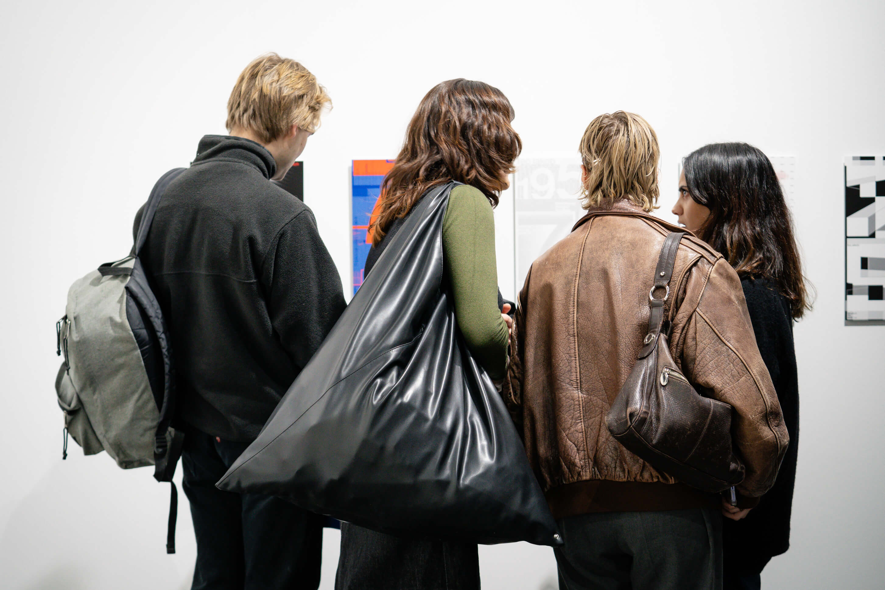 Vier Personen stehen mit dem Rücken zur Kamera in einer Ausstellung und betrachten grafische Arbeiten an einer weissen Wand, zwei tragen grosse Taschen und Jacken.