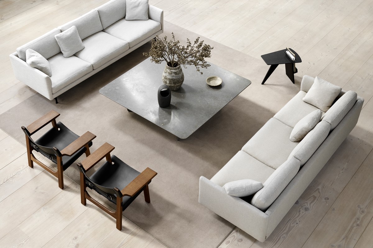 Draufsicht auf eine helle Wohnzimmergruppe mit zwei beigen Sofas und zwei Holz Sesseln mit schwarzer Bespannung. In der Mitte steht ein grosser, quadratischer Couchtisch mit Vase und Schalen auf einem hellen Teppich. Rechts ein kleiner schwarzer Beistelltisch, rundherum heller Holzboden.