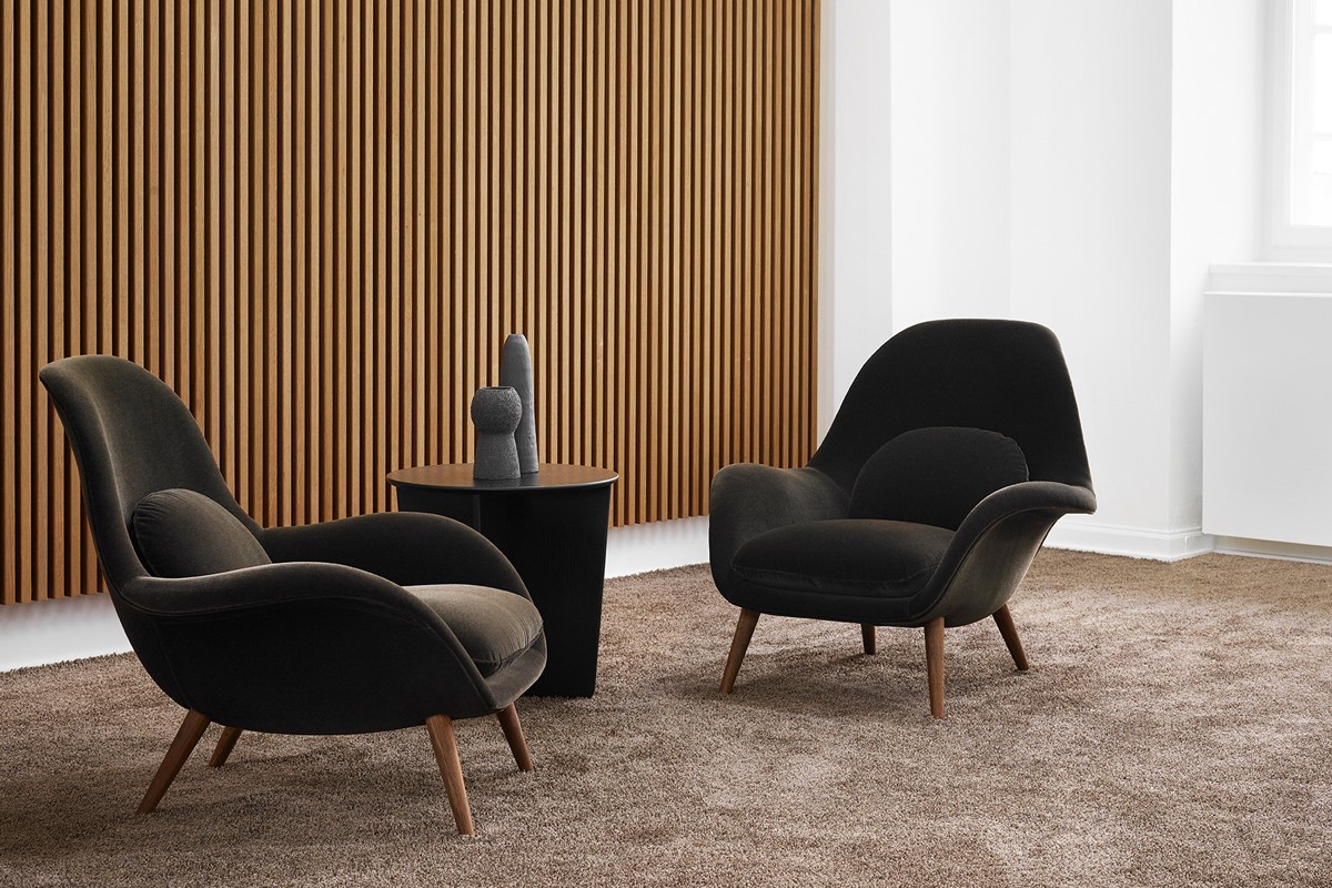 Zwei schwarze Lounge Sessel mit Holzfüssen stehen auf einem braunen Teppich. Dazwischen ein runder, dunkler Beistelltisch mit zwei skulpturalen Vasen. Im Hintergrund eine Wand mit vertikalen Holzlamellen, rechts eine helle Wandzone.