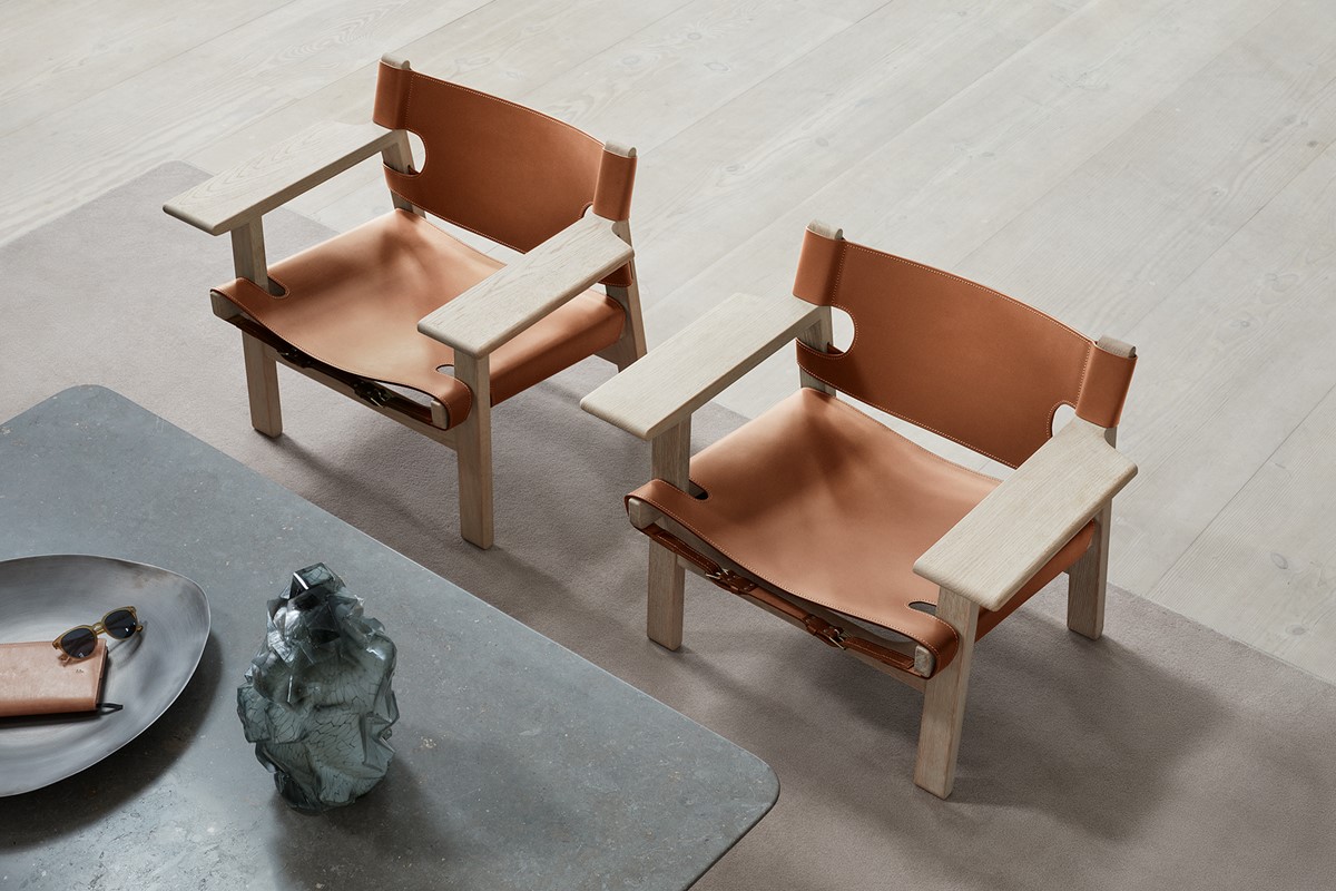 Zwei Lounge Sessel mit Gestell aus hellem Holz und Sitzflächen aus cognacfarbenem Leder stehen auf einem hellen Teppich. Im Vordergrund ist ein Teil eines niedrigen Tisches mit Schale, Sonnenbrille und Dekoobjekt zu sehen. Heller Holzboden und ruhige, minimalistische Umgebung.