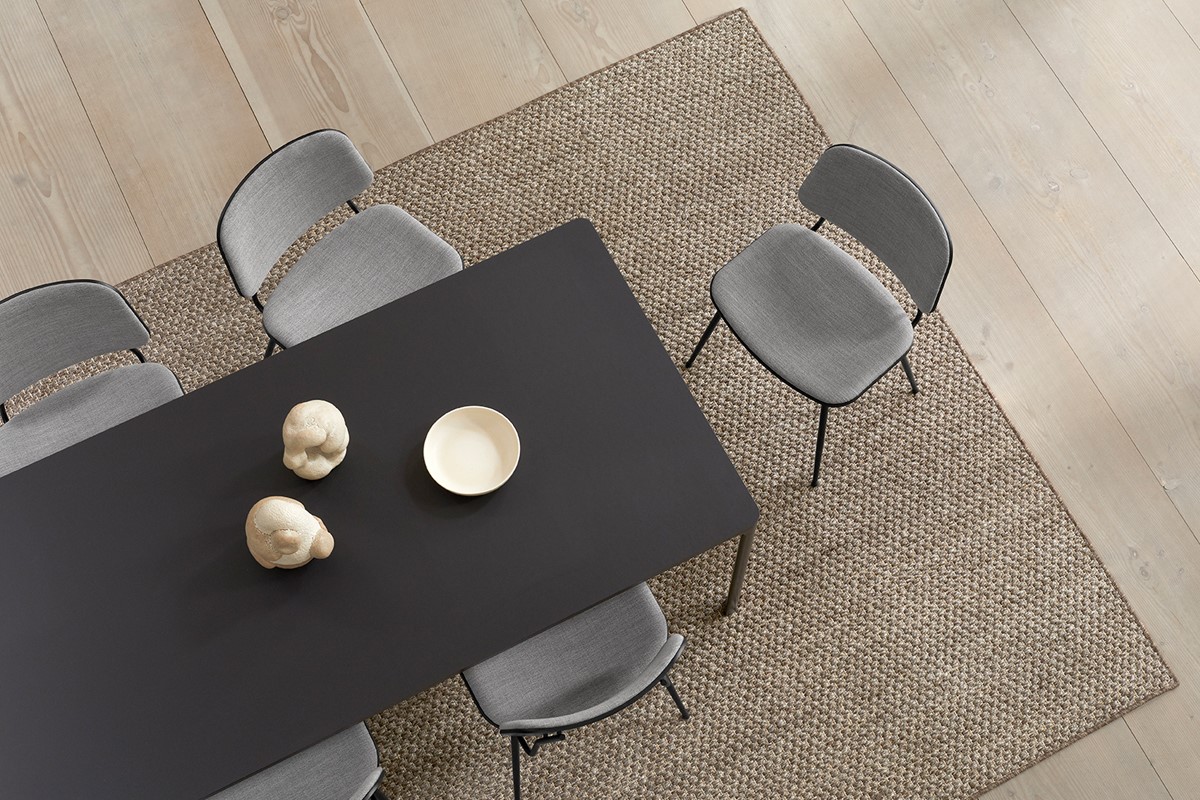 Draufsicht auf einen schwarzen Esstisch auf grob gewebtem Naturteppich. Rundum stehen graue Polsterstühle mit schwarzem Gestell. Auf dem Tisch liegen zwei helle Dekoobjekte und eine weisse Schale. Heller Holzboden rahmt das Setting.