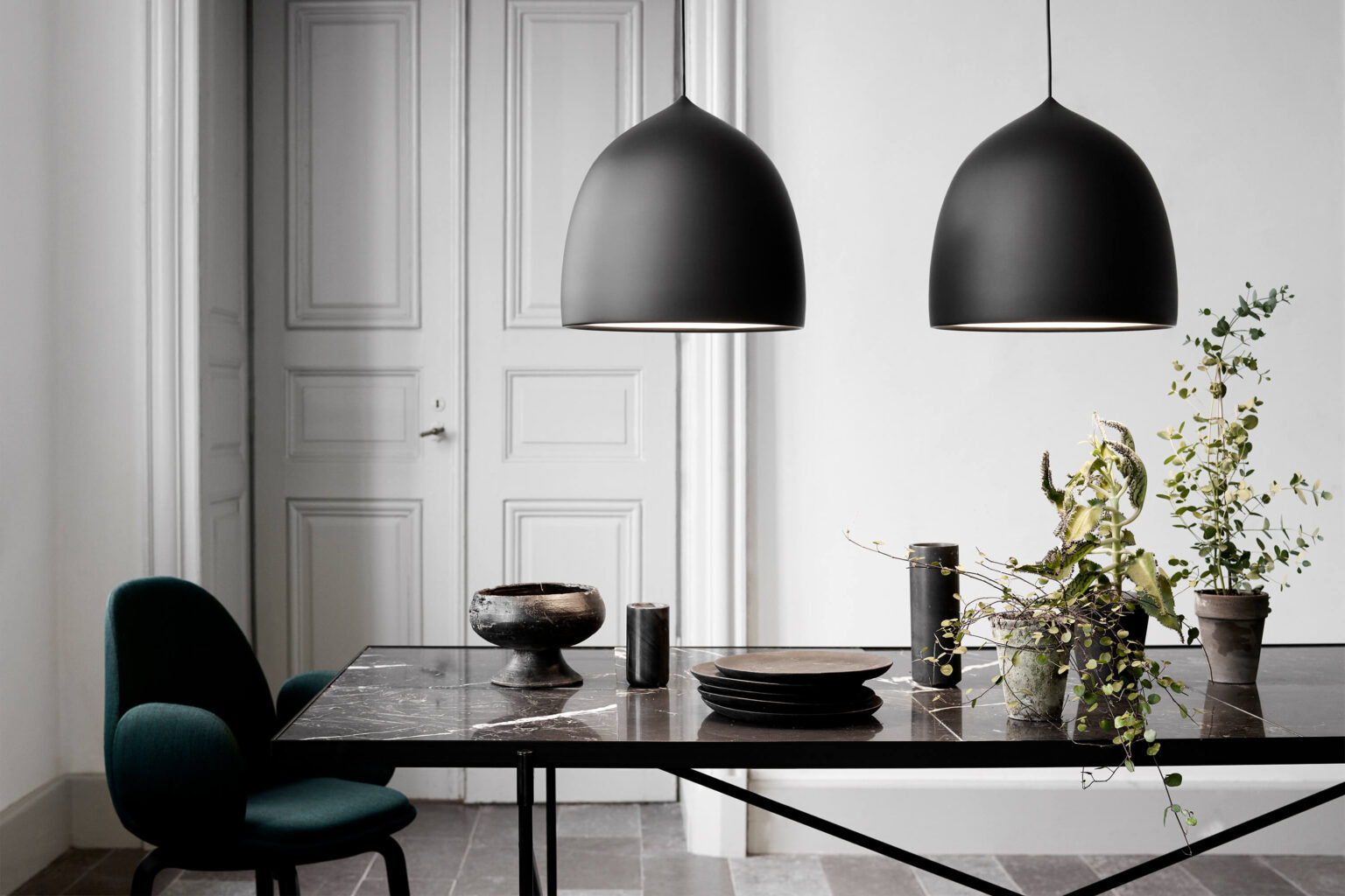 Essbereich mit schwarzem Tisch in Steinoptik und dunklem Stuhl. Über dem Tisch hängen zwei grosse, matte Pendelleuchten in Schwarz. Auf der Tischplatte stehen Keramikgeschirr, Vasen und ein Arrangement aus grünen Zweigen. Im Hintergrund eine helle Wand mit klassischer Tür.