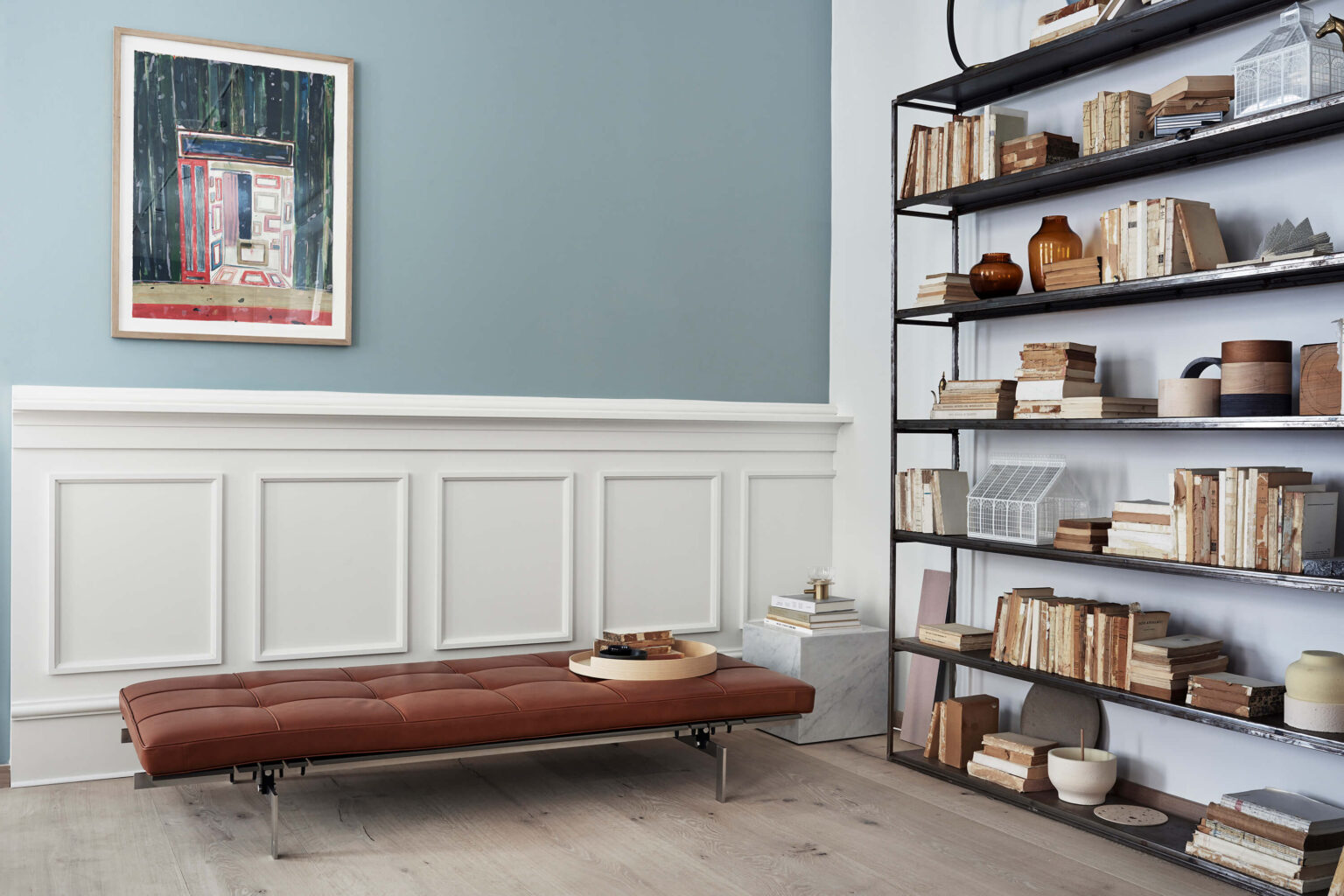 Wohnraum mit brauner, gepolsterter Liege auf hellem Holzboden. Links eine Wand mit weisser Täfelung und blauer Fläche, darüber ein gerahmtes Kunstwerk. Rechts ein hohes Metallregal mit vielen Büchern und Keramikobjekten. Neben der Liege steht ein kleiner Beistelltisch mit Tablett und Büchern.
