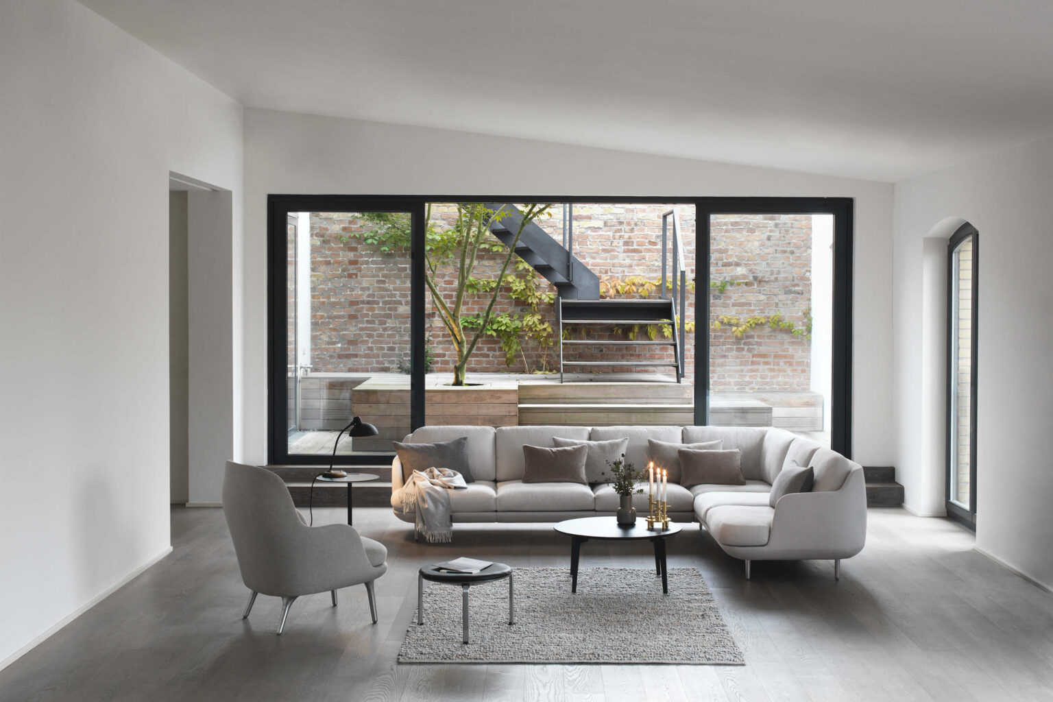 Modernes Wohnzimmer mit hellem Ecksofa vor einer grossen Fensterfront. Davor stehen runde Couchtische mit Kerzen und Deko auf einem strukturierten Teppich. Links ein Sessel und Beistelltisch, rechts viel freie Fläche. Draussen sind eine Backsteinwand, Pflanzen und eine Metalltreppe im Innenhof zu sehen.