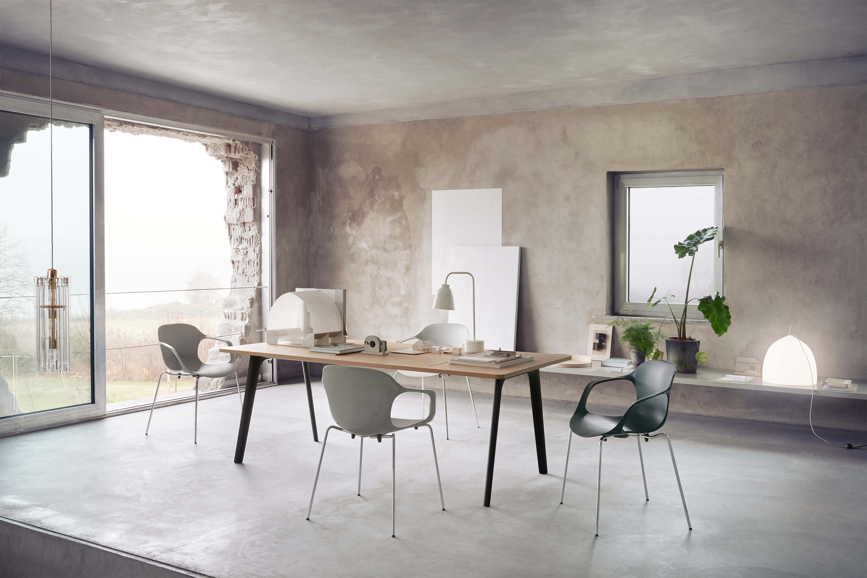 Heller Arbeitsraum mit rohen, grauen Wänden und Betonboden. In der Mitte steht ein langer Holztisch mit Modellobjekten, Papieren und einer kleinen Tischleuchte. Rundum stehen leichte Stühle mit Metallgestell. Links öffnet eine grosse Glasfront den Blick ins Grüne, rechts eine Fensterbank mit Pflanzen und leuchtender Tischlampe.