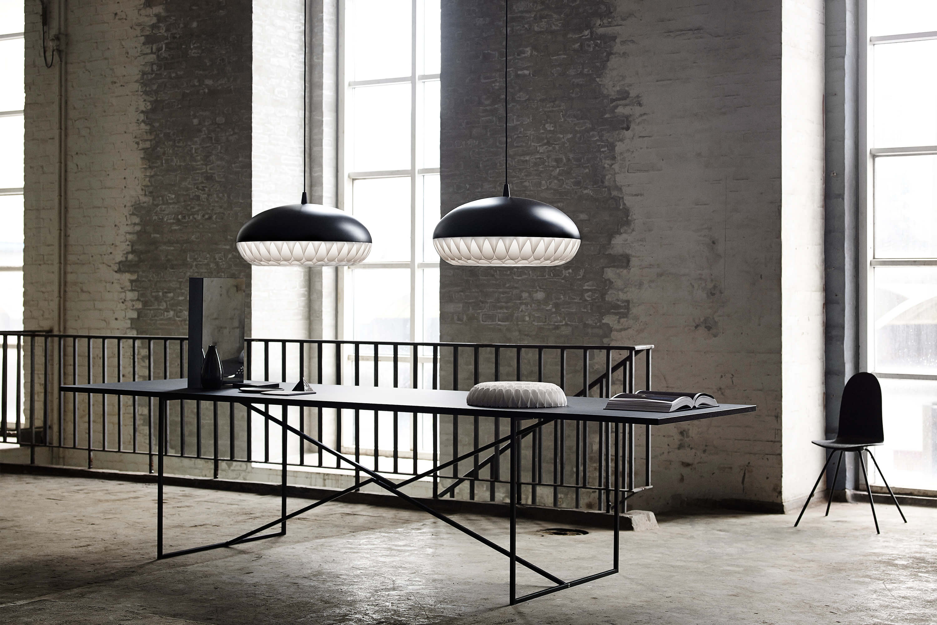 Industrie Loft mit rauen Backsteinwänden und hohen Fenstern. Über einem langen, schwarzen Tisch hängen zwei grosse Pendelleuchten mit weissem Diffusor. Auf dem Tisch liegen Vasen, ein Kissen und ein aufgeschlagenes Buch. Rechts steht ein einzelner schwarzer Stuhl, im Hintergrund ein Metallgeländer.