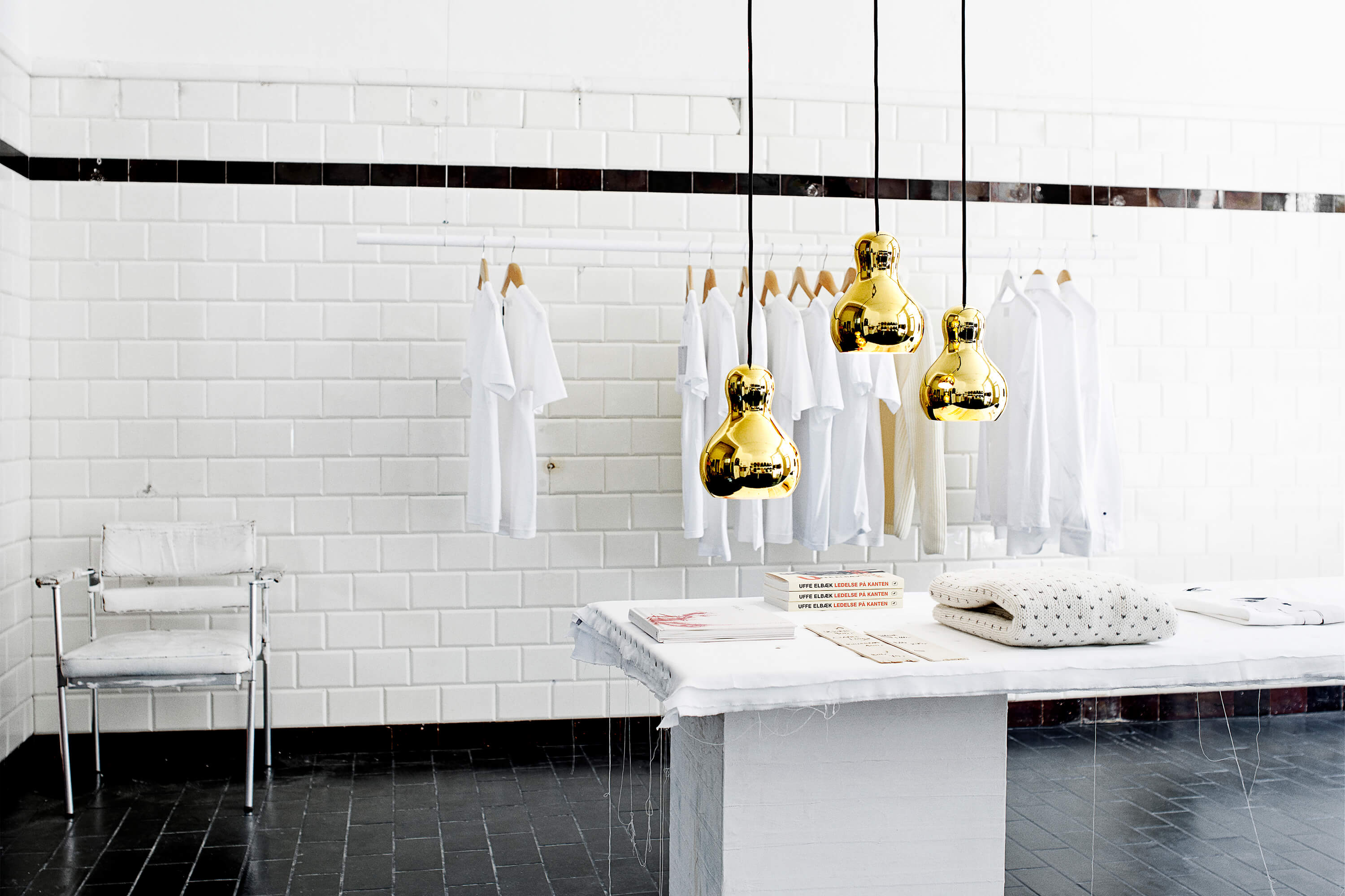 Heller Boutique Raum mit weisser Fliesenwand und Kleiderstange mit weissen Shirts und Hemden. Über einem zentralen Tisch hängen drei goldene Pendelleuchten mit spiegelnder Oberfläche. Auf dem Tisch liegen Bücher und gefaltete Textilien. Links steht ein transparenter Stuhl, der Boden ist dunkel gefliest.