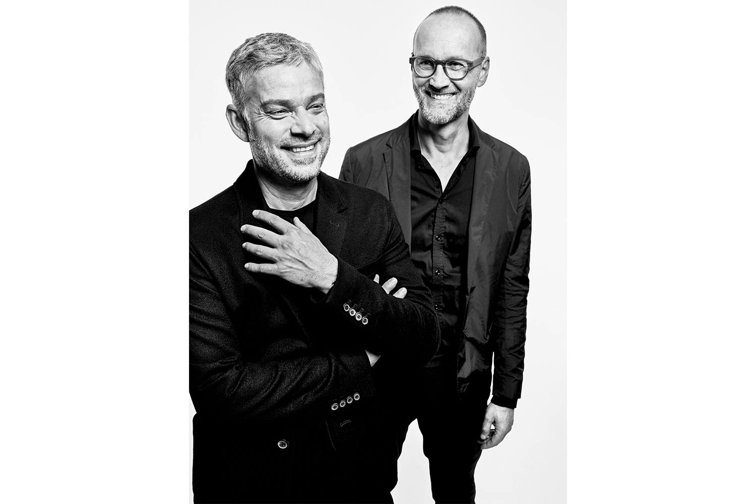 Schwarzweisses Studio-Porträt von zwei lachenden Männern in dunkler Kleidung. Vorn links ein Mann mit kurzem grauem Haar und Bart, Hand an der Brust. Hinten rechts ein Mann mit Brille und kurzem Bart.