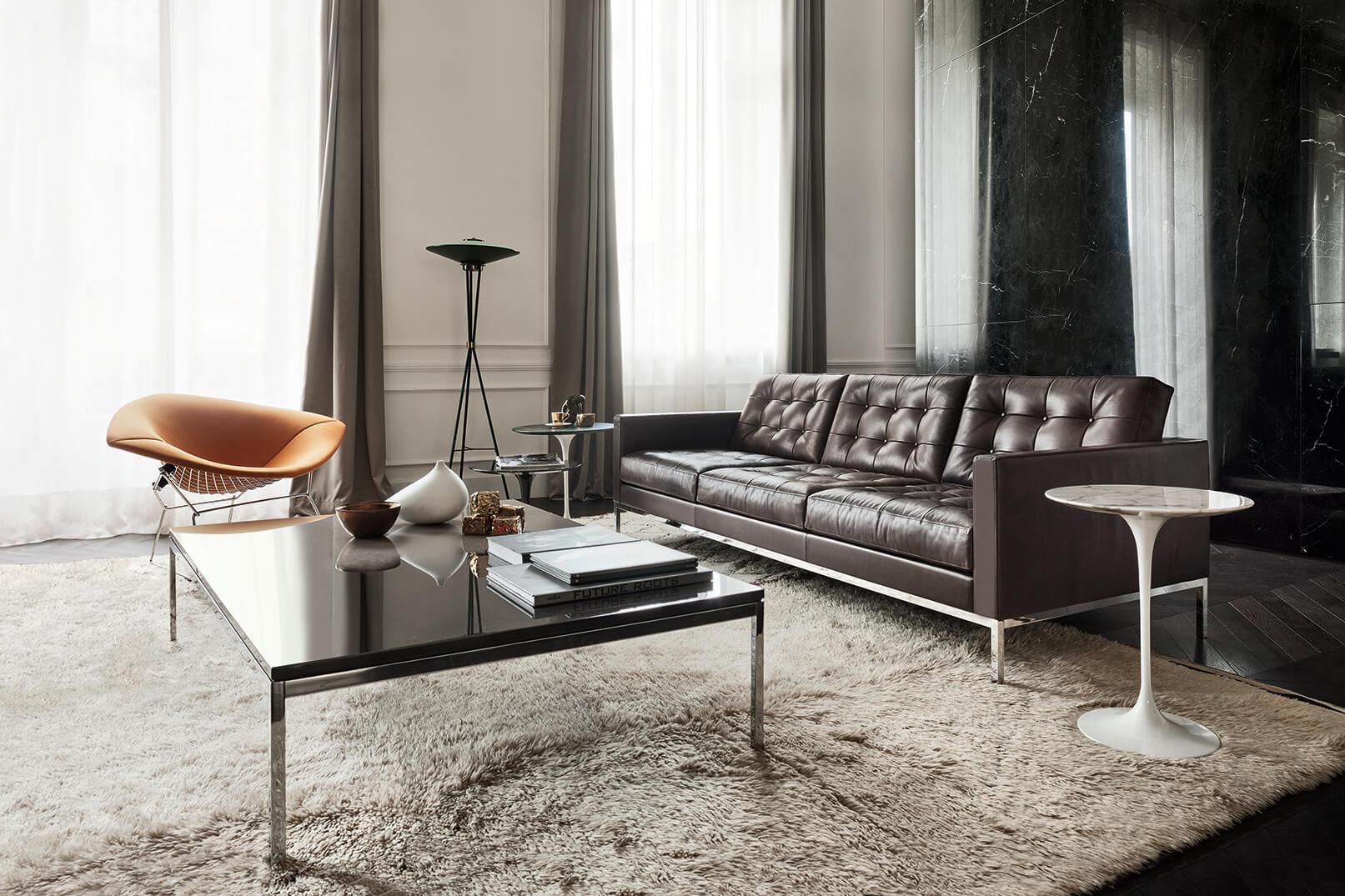Elegantes Wohnzimmer mit hoher Fensterfront und bodenlangen Vorhängen. Rechts steht ein dunkelbraunes, gestepptes Ledersofa vor einer schwarzen Marmorwand. Davor ein grosser, flauschiger Teppich mit schwarzem Glastisch und Dekorobjekten. Links ein orangener Loungesessel, im Raum eine schlanke Stehleuchte und ein kleiner weisser Beistelltisch.