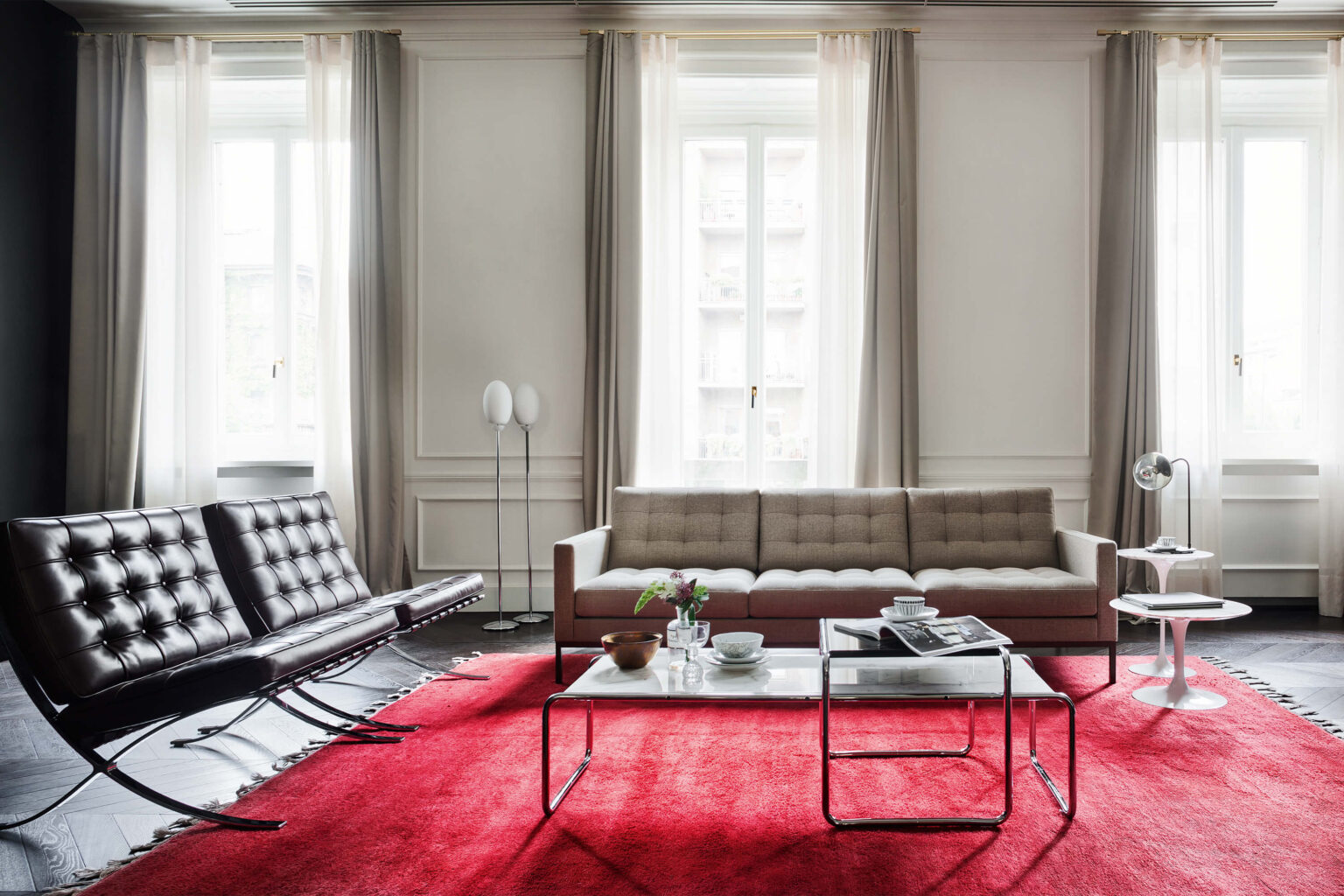 Helles Wohnzimmer mit hohen Fenstern und hellen Vorhängen. In der Mitte steht ein beiges Sofa auf einem grossen roten Teppich, davor ein niedriger Couchtisch mit Geschirr und Zeitschriften. Links eine schwarze Lederliege, rechts ein kleiner runder Beistelltisch mit Tischleuchte. Dezente Stehleuchten und klassische Wandpaneele im Hintergrund.