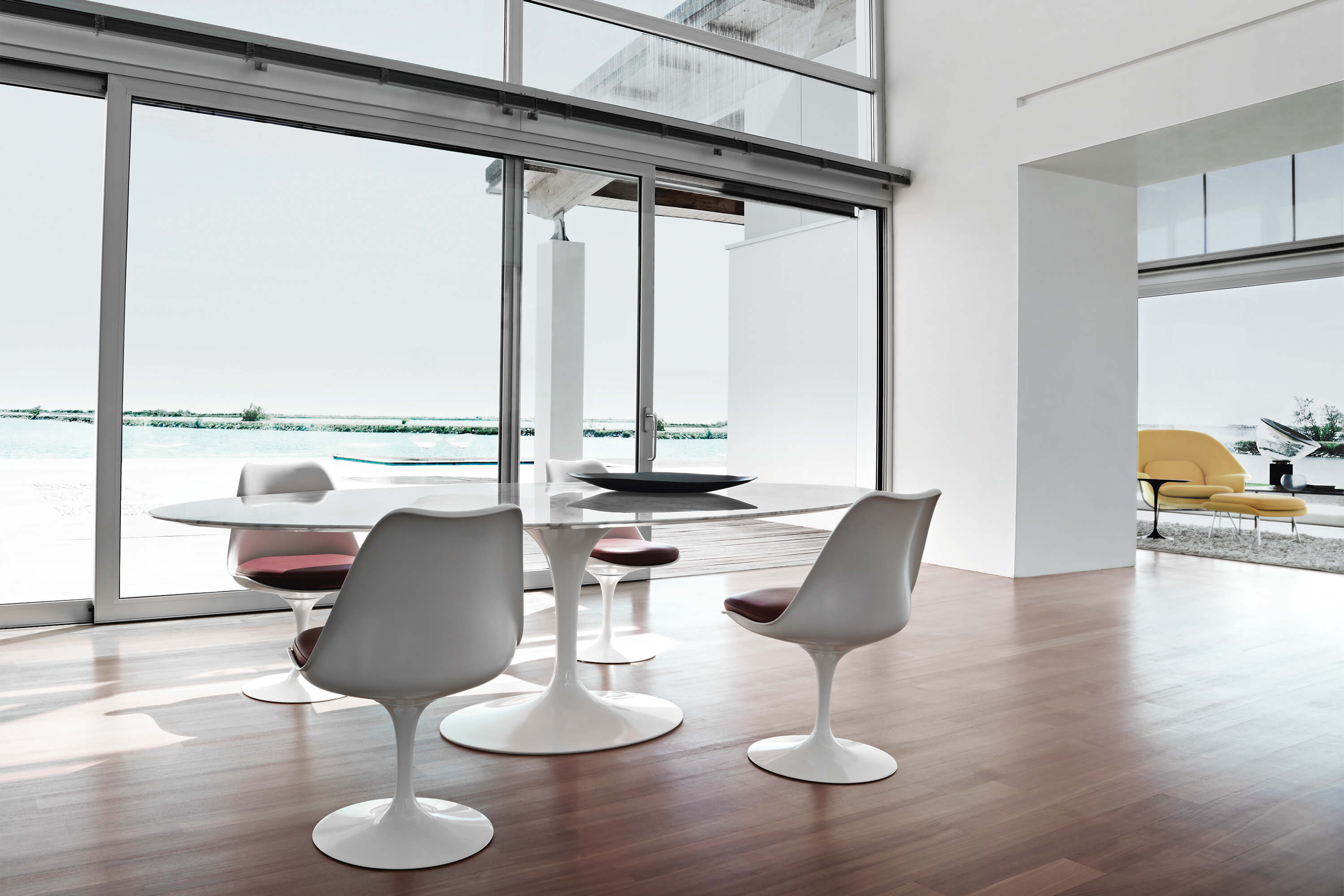 Lichtdurchfluteter Essbereich mit rundem weissem Tulip Tisch und vier weissen Tulip Stühlen, bodentiefen Schiebetüren und Blick auf Wasser und Terrasse, im Hintergrund ein angrenzender Raum mit gelbem Loungesessel.