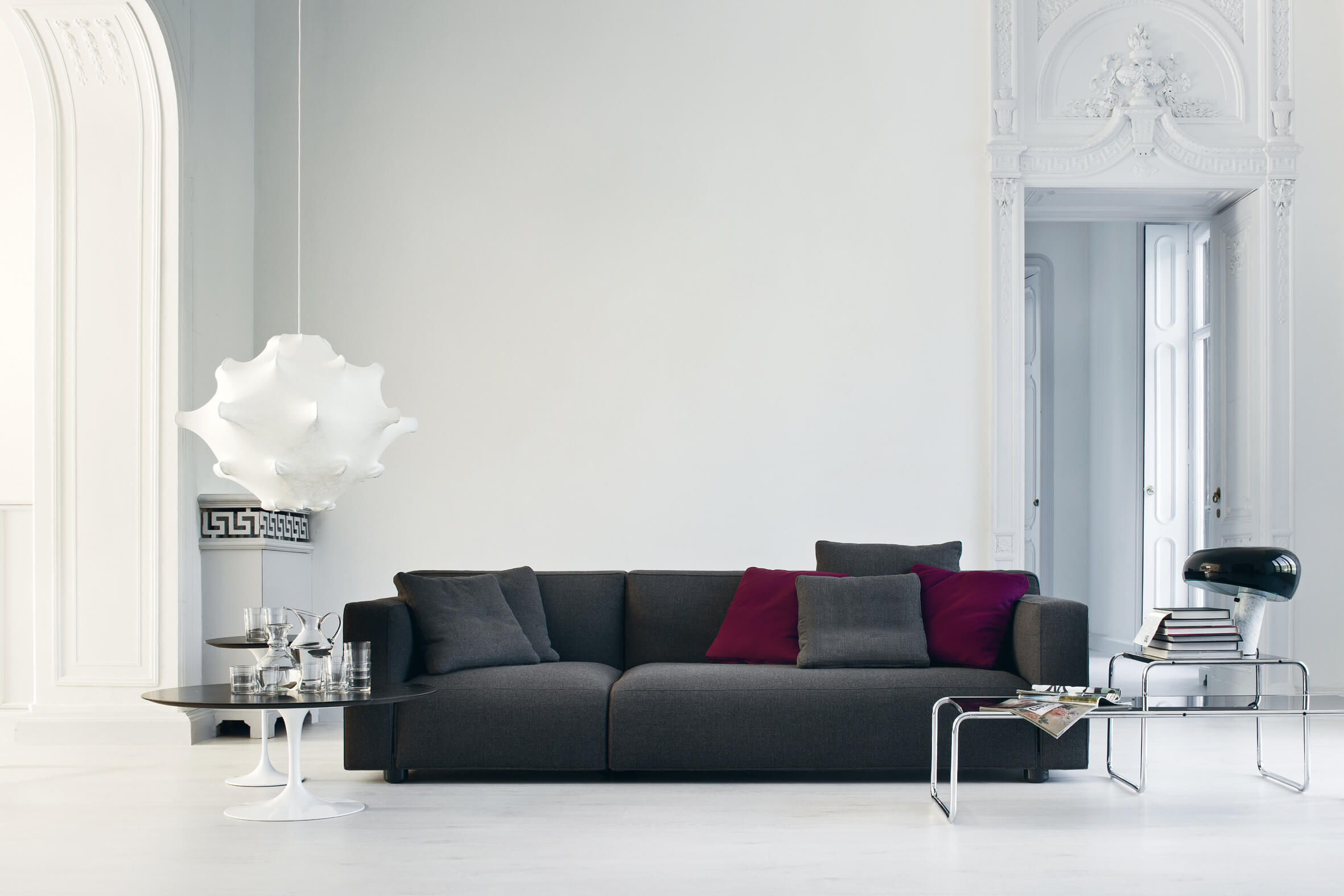 Helles, klassisches Wohnzimmer mit dunkelgrauem Sofa und bordeauxroten Kissen, skulpturaler weisser Pendelleuchte sowie Beistelltischen und Chrom Couchtisch vor dekorativer Stucktüre.