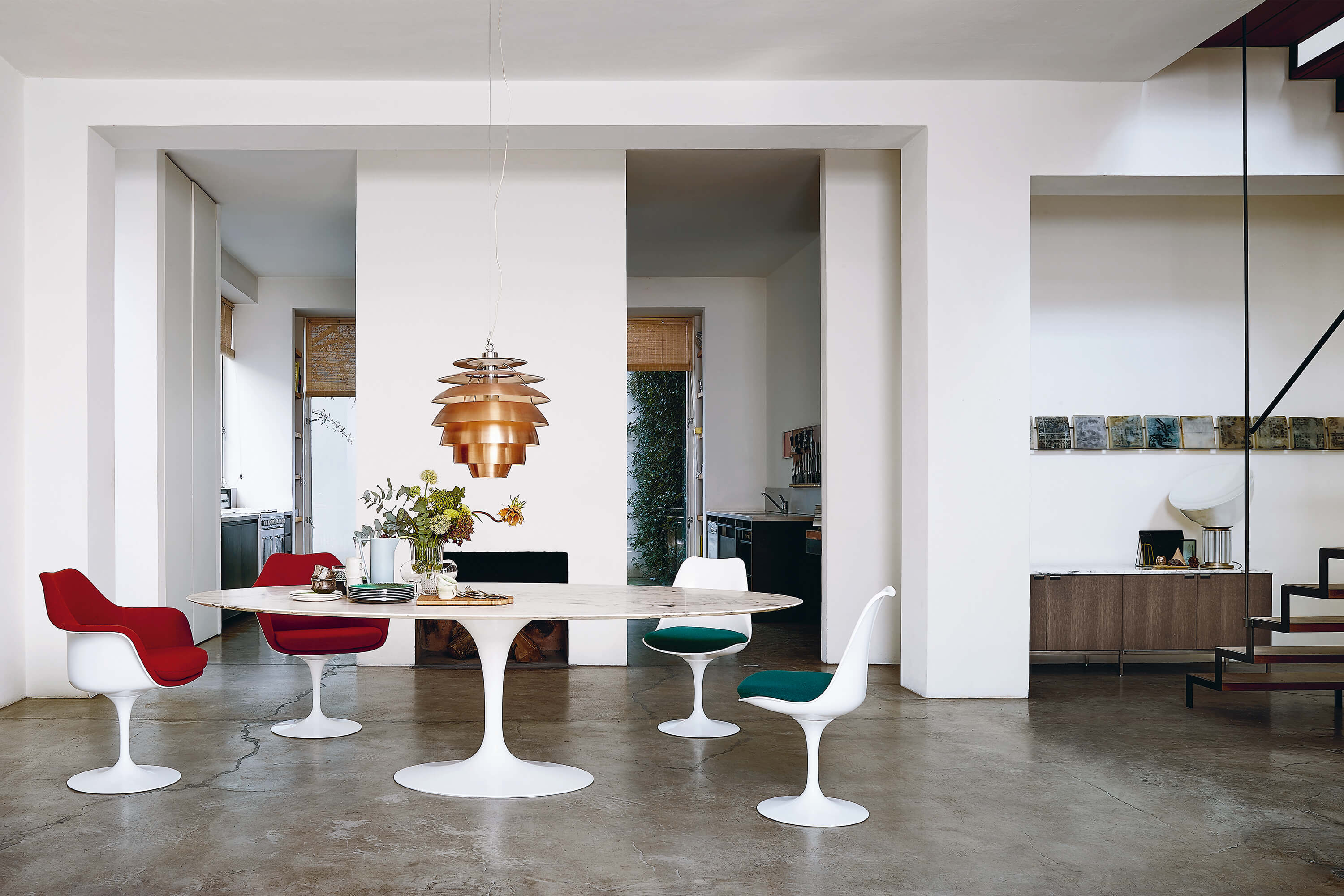 Minimalistischer Essbereich mit weissem Tulip Tisch auf Sichtbetonboden. Rundherum stehen Tulip Stühle in Rot, Weiss und Grün, darüber hängt eine kupferfarbene Pendelleuchte, im Hintergrund weisse Durchgänge und ein Sideboard mit Objekten.