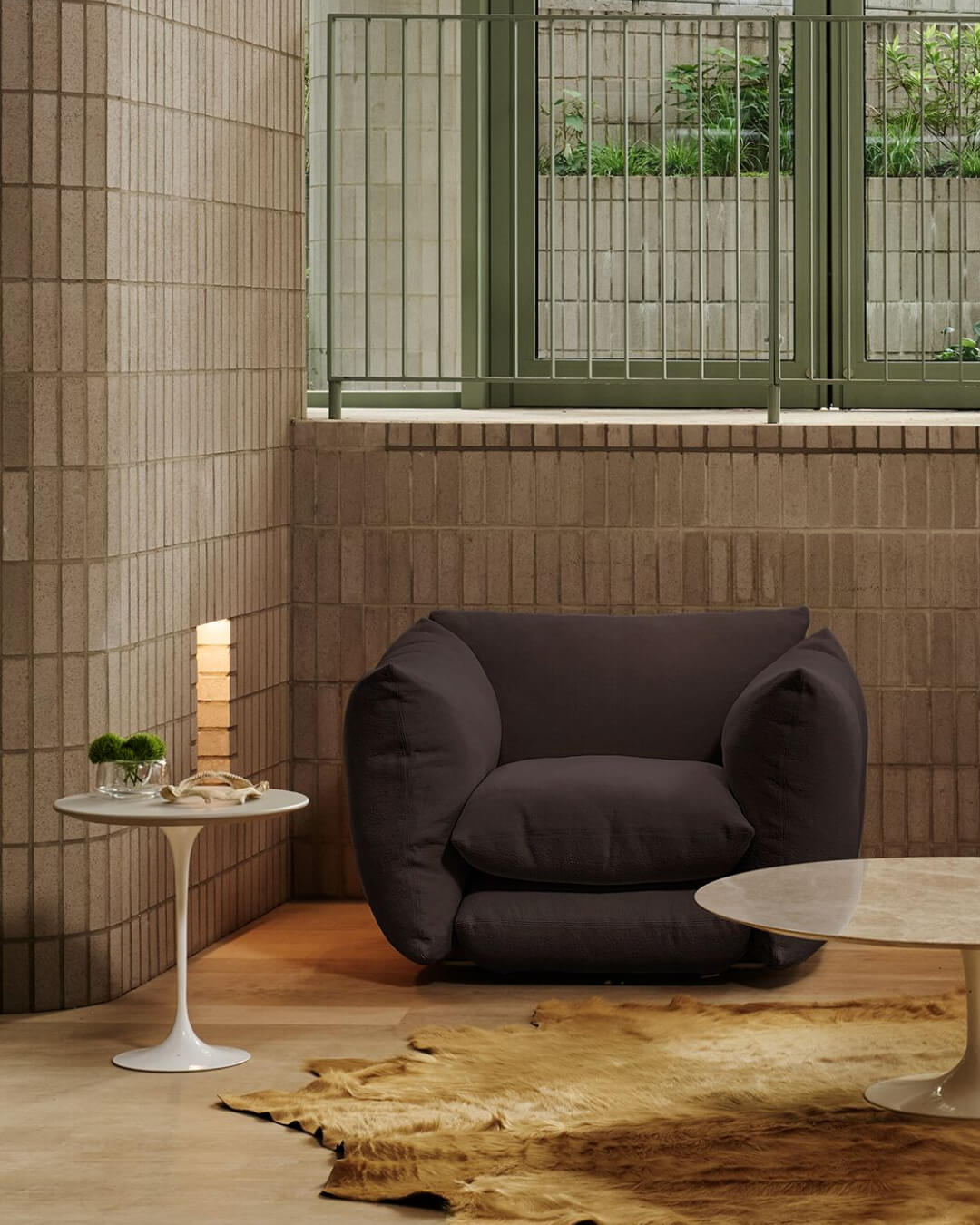 Ruhige Lounge Ecke mit grosszügigem, dunkelgrauem Sessel vor hellen Klinkerwänden. Links ein weisser Beistelltisch und eine niedrige Leuchte, vorne ein heller Marmortisch und ein Fellteppich, dahinter ein grosses Fenster mit Blick ins Grüne.