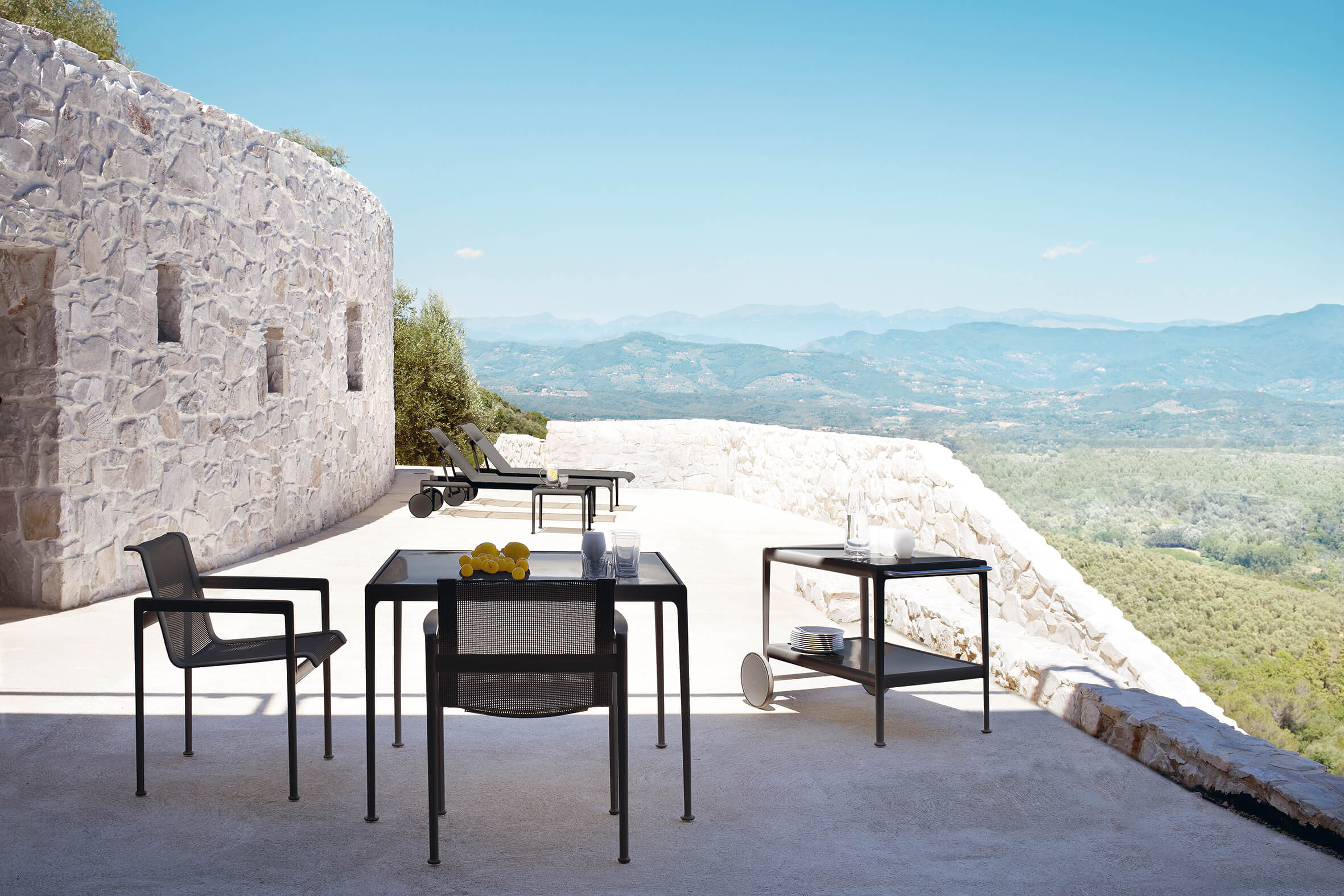 Helle Steinterrasse mit schwarzem Outdoor Esstisch und Stühlen. Rechts ein Servierwagen mit Geschirr, im Hintergrund zwei Liegen. Weite Aussicht über grüne Hügel bis zu fernen Bergen unter blauem Himmel.