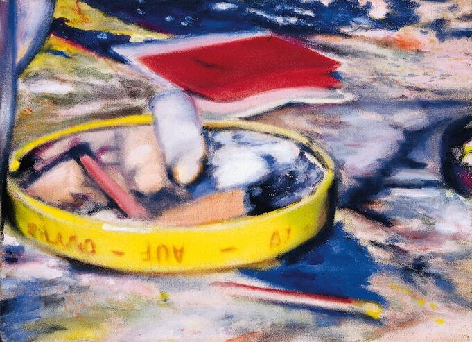 Unscharfe Atelier-Szene in kräftigen Farben. Ein gelber runder Deckel dient als Schale mit Stiften und kleinen Objekten. Daneben liegt ein rotes Heft auf einem farbverschmierten Tisch in Blau, Gelb und Rosa.