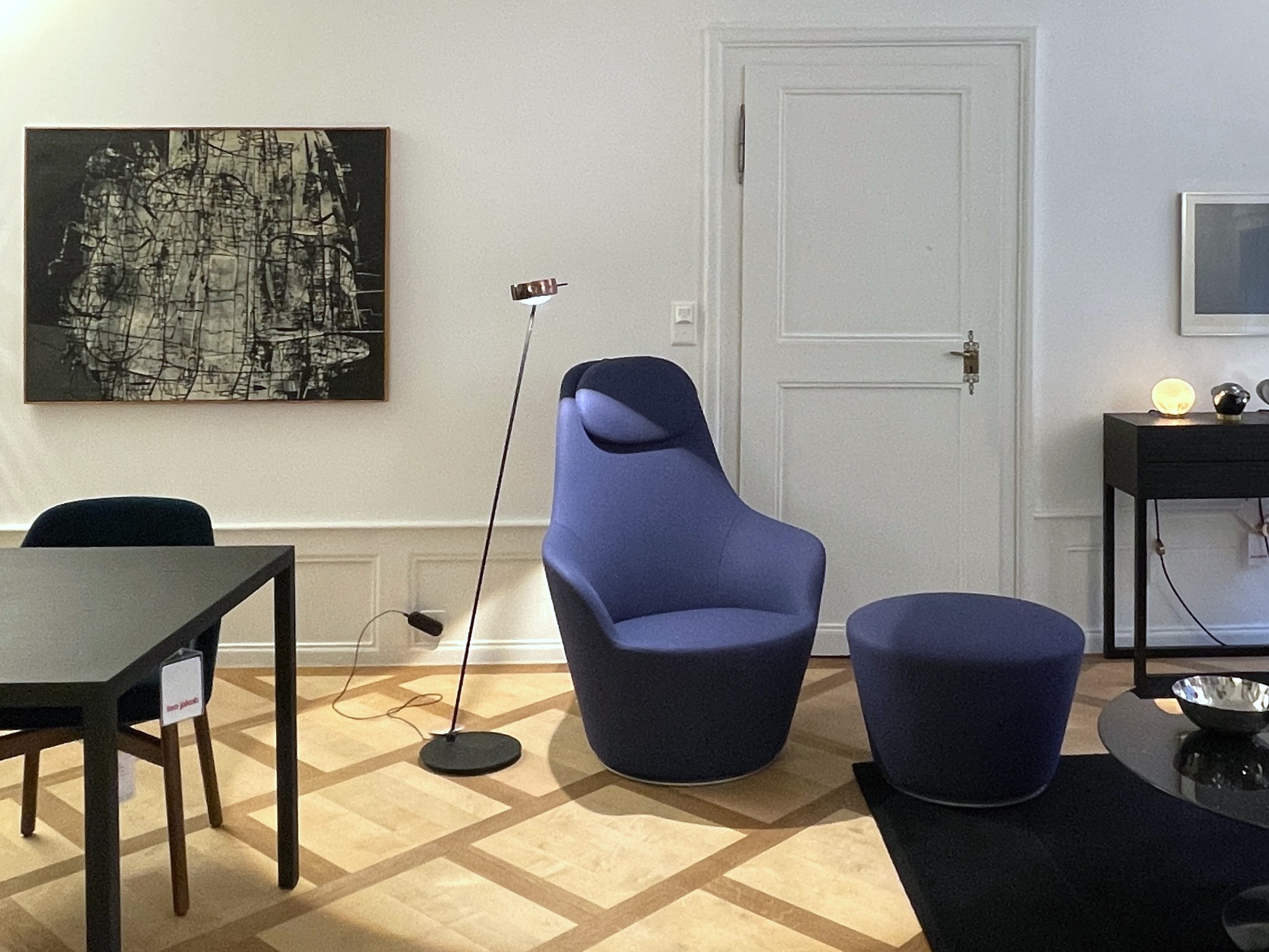 Wohnszene mit blauem Lounge-Sessel und passendem Hocker neben einer schlanken Stehleuchte. Links hängt ein abstraktes Schwarz-Grau-Bild, rechts Tür und schwarze Konsole mit Leuchten, Parkettboden und schwarzer Esstisch im Vordergrund.