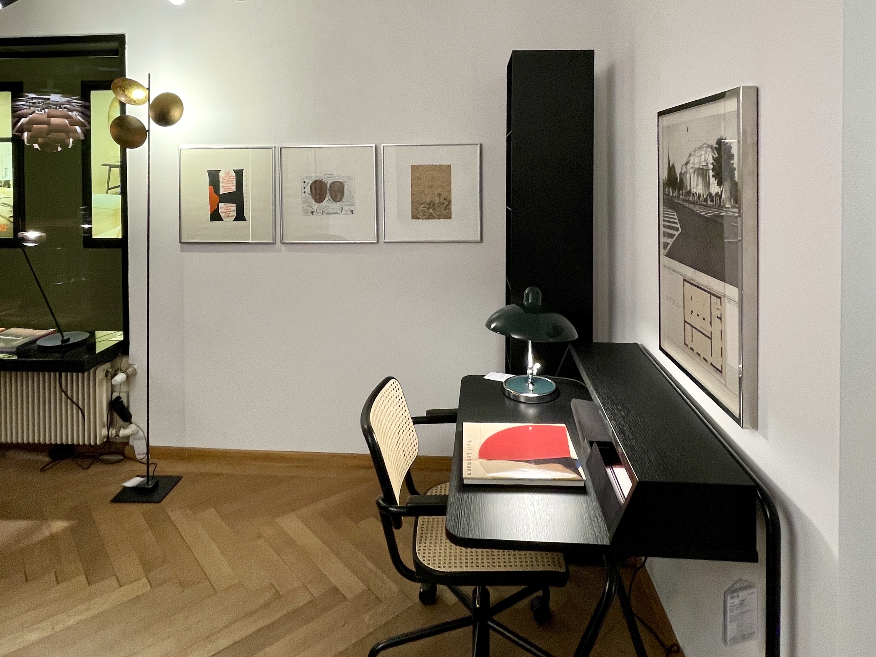 Stilvolle Arbeitsecke mit schwarzem Sekretär und Stuhl mit Rohrgeflecht, darauf eine grüne Tischlampe und ein aufgelegtes Buch. Rechts hängt eine großformatige, gerahmte Visualisierung einer verhüllten Kunsthalle, links drei weitere Grafiken und eine skulpturale Stehleuchte. Parkettboden, Fensterzone links, ruhige Galerieatmosphäre.