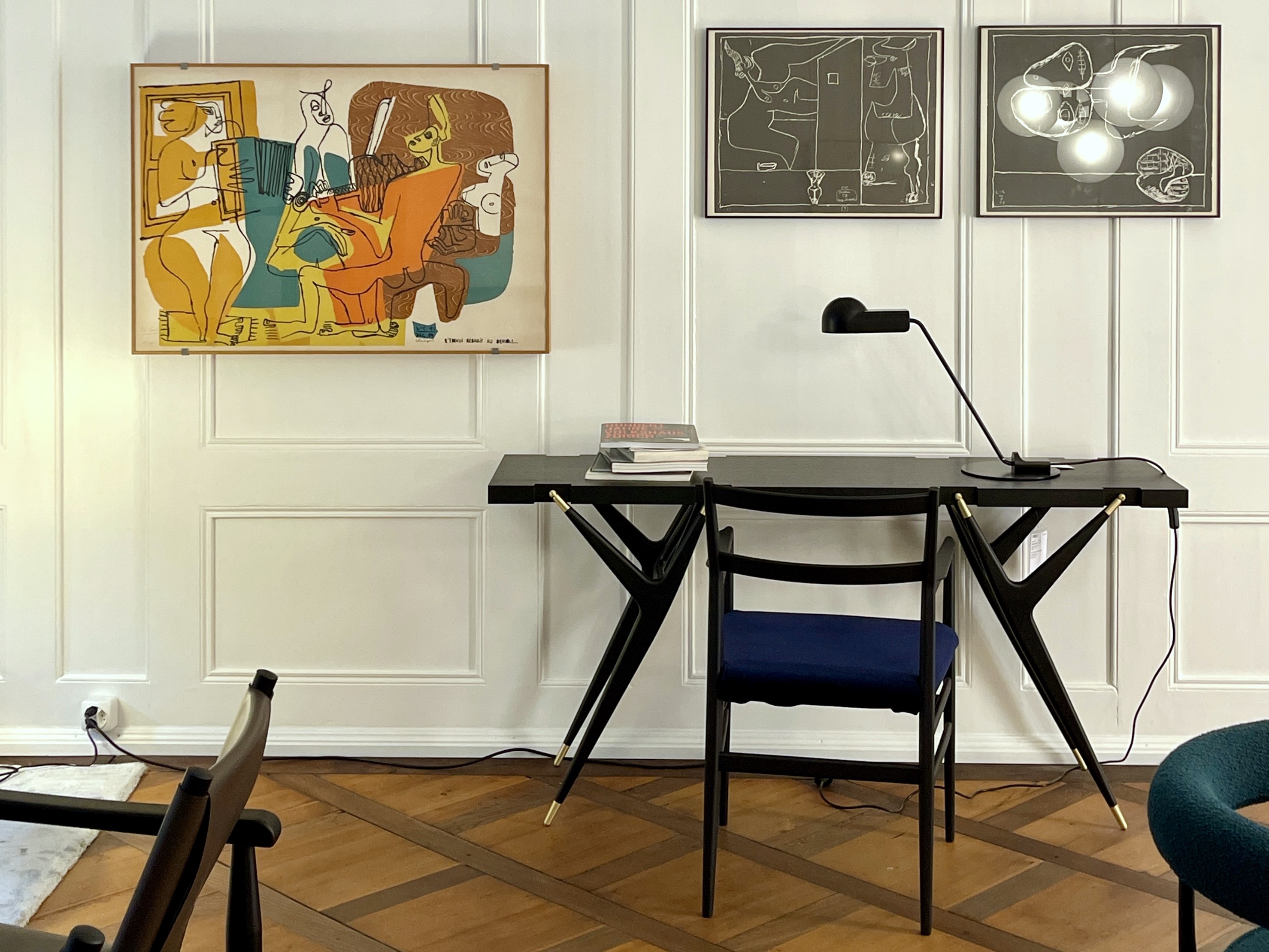 Heller Raum mit Wandvertäfelung und Parkett. Schwarzer Schreibtisch mit filigranen V-Beinen, dazu ein Stuhl mit blauem Sitz und eine schwarze Tischleuchte. An der Wand hängen drei gerahmte Grafiken, links eine farbige Komposition, rechts zwei monochrome Blätter. Auf dem Tisch liegen zwei Bücher.