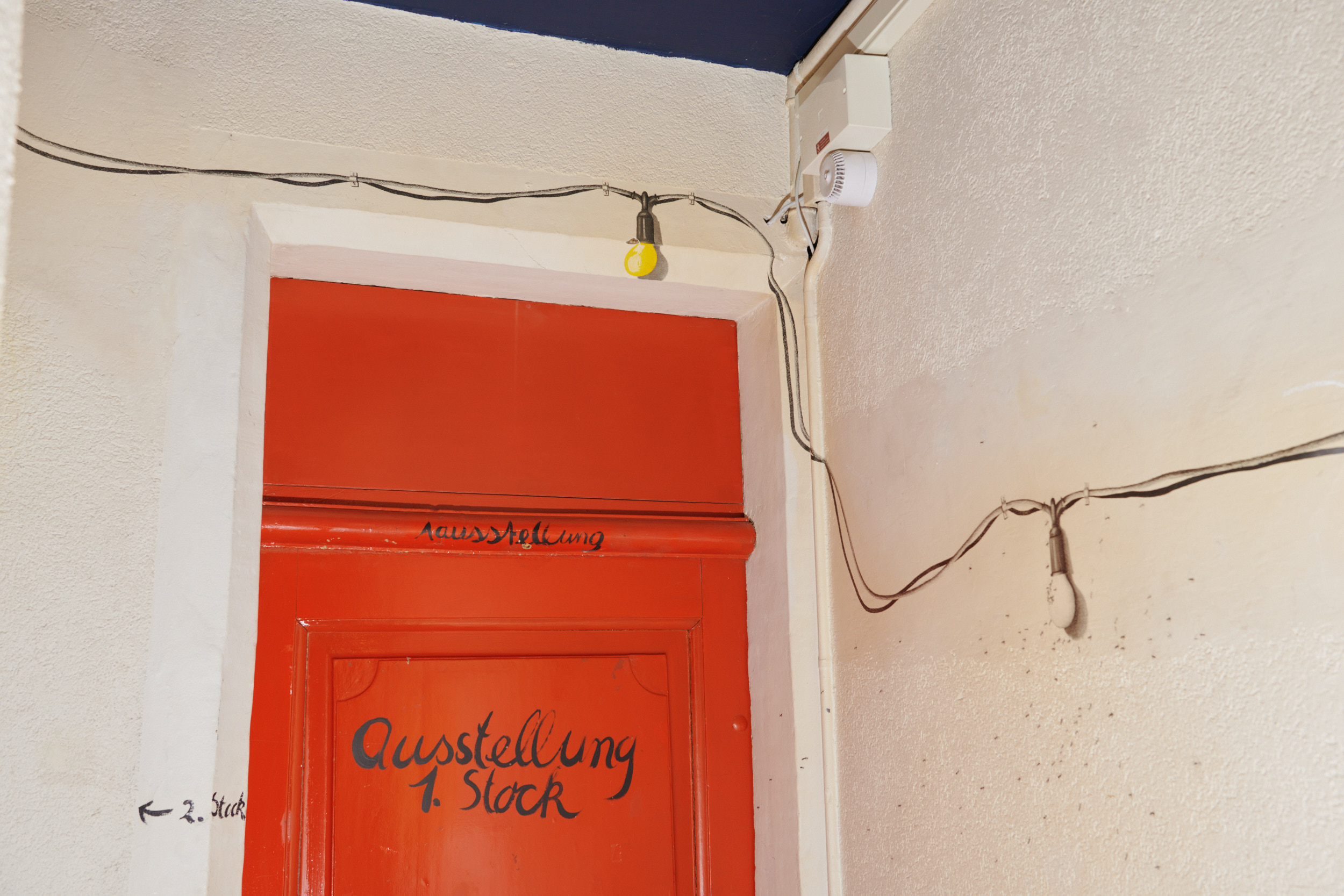 Rote Tür im Treppenhaus mit der Aufschrift „Ausstellung 1. Stock“. Handgeschriebener Pfeil weist zum „2. Stock“. Sichtbare Kabel und nackte Glühlampen an heller, rauer Wand. Rohes, improvisiertes Ambiente.