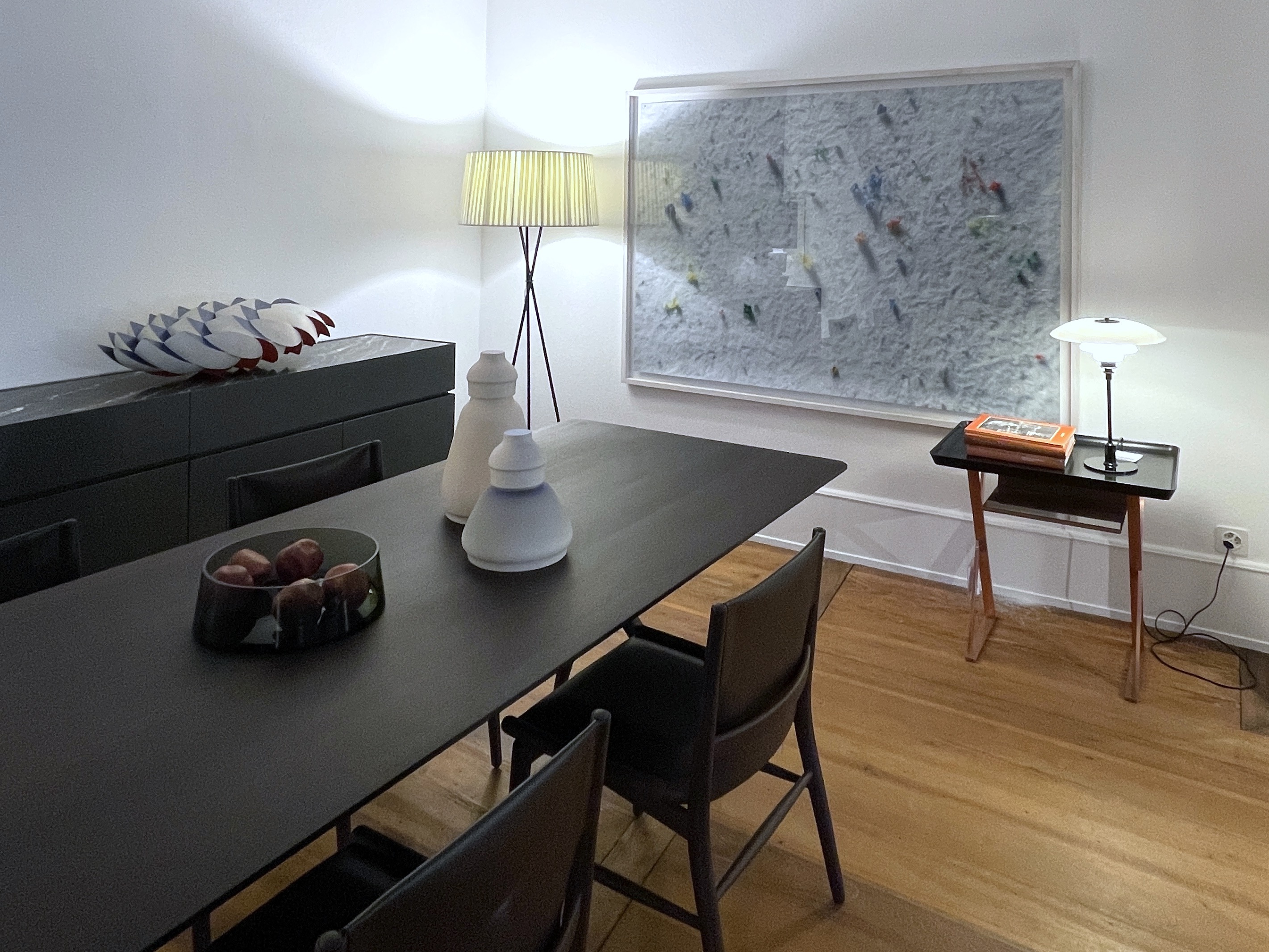 Heller Essbereich mit schwarzem Tisch und dunklen Stühlen auf Holzboden. An der Wand hängt ein grosses, gerahmtes Kunstwerk mit farbigen Partikeln, flankiert von Stehleuchte und Beistelltisch mit Tischlampe; links steht ein Sideboard mit skulpturalem Objekt.