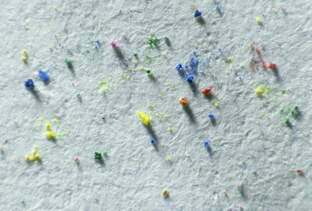 Makroaufnahme einer hellen Papieroberfläche mit verstreuten Farbpartikeln in Blau, Gelb, Rot und Grün. Feine Fasern und Schatten zeigen die Struktur des Papiers.