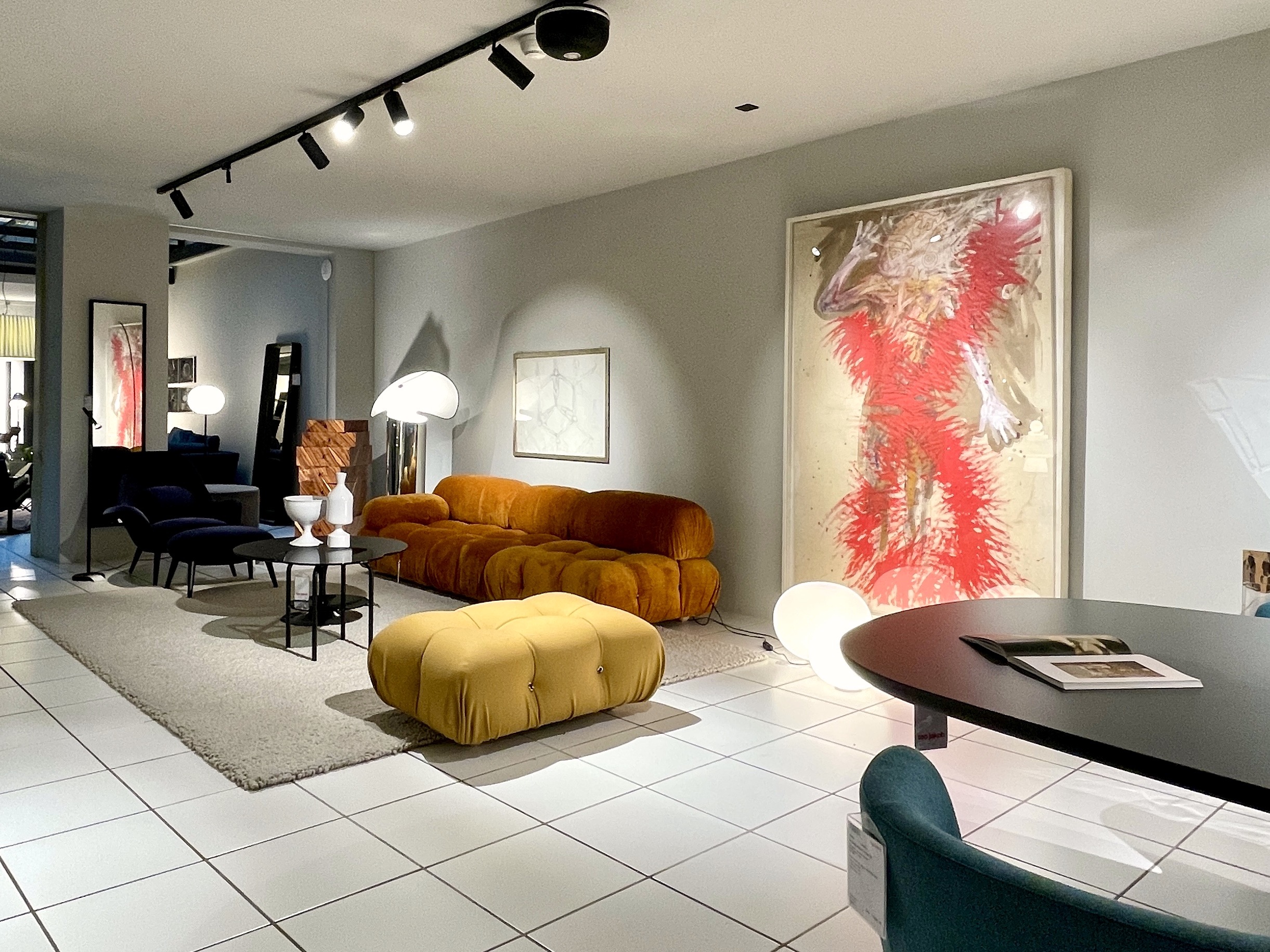 Design-Showroom mit ockerfarbenem Modularsofa und passendem Pouf auf hellem Teppich. An der grauen Wand hängt ein grosses Bild mit roter Figur, dazu runde Leuchten, Couchtische und ein schwarzer runder Tisch im Vordergrund.