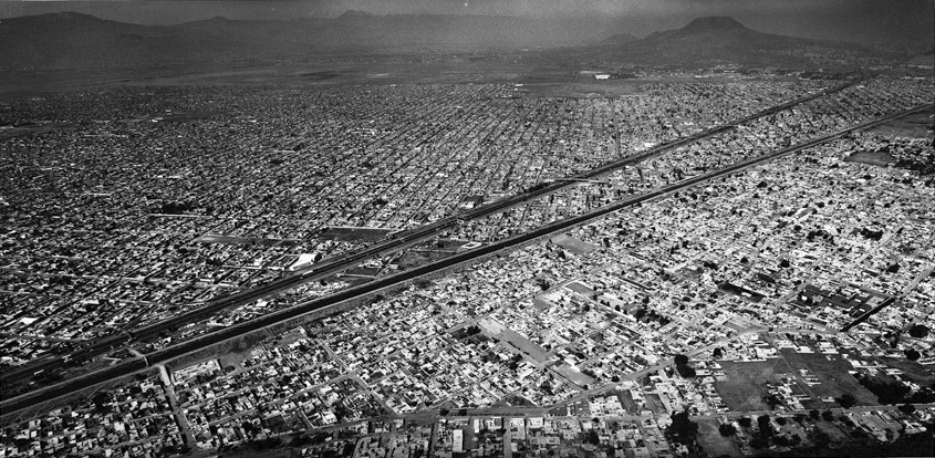 Schwarzweisse Luftaufnahme einer ausgedehnten Stadtlandschaft. Eine breite Schnellstrasse verläuft diagonal durch das dichte Häusermeer, im Hintergrund Berge im Dunst.