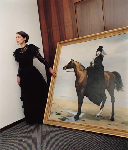 Frau in schwarzem Kleid steht in einem Innenraum neben einem grossen, gerahmten Gemälde. Das Bild zeigt eine elegant gekleidete Reiterin seitlich auf einem braunen Pferd am Strand.