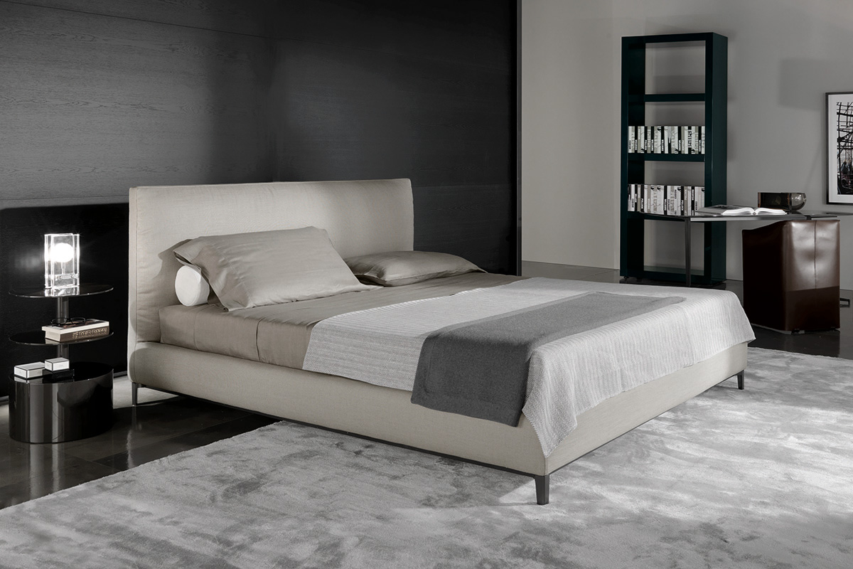 Modernes Schlafzimmer mit gepolstertem Bett in hellem Grau, grauer Bettwäsche und Plaid auf grossem Teppich, dazu Nachttisch mit Glasleuchte, schwarzes Regal mit Büchern und Schreibtisch im Hintergrund.