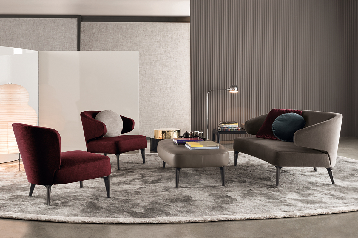 Elegante Lounge mit grauem Teppich, zwei bordeauxfarbenen Sesseln und grauem Sofa um einen ovalen Couchtisch, ergänzt durch Beistelltisch mit Leuchte und strukturierte Wandpaneele.