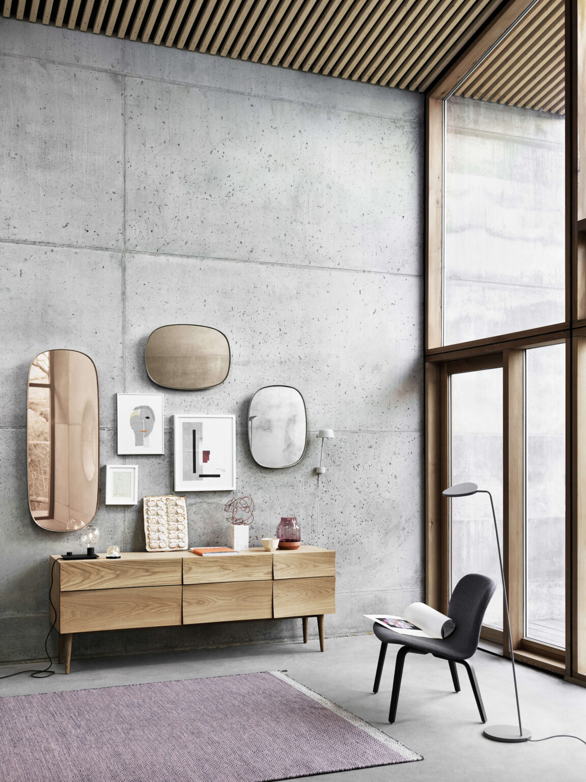 Modernes Interieur mit Sichtbetonwand, Holz Sideboard mit Deko, Spiegeln und gerahmten Grafiken, dazu Loungesessel und Stehleuchte neben bodentiefem Fenster mit Holzrahmen.