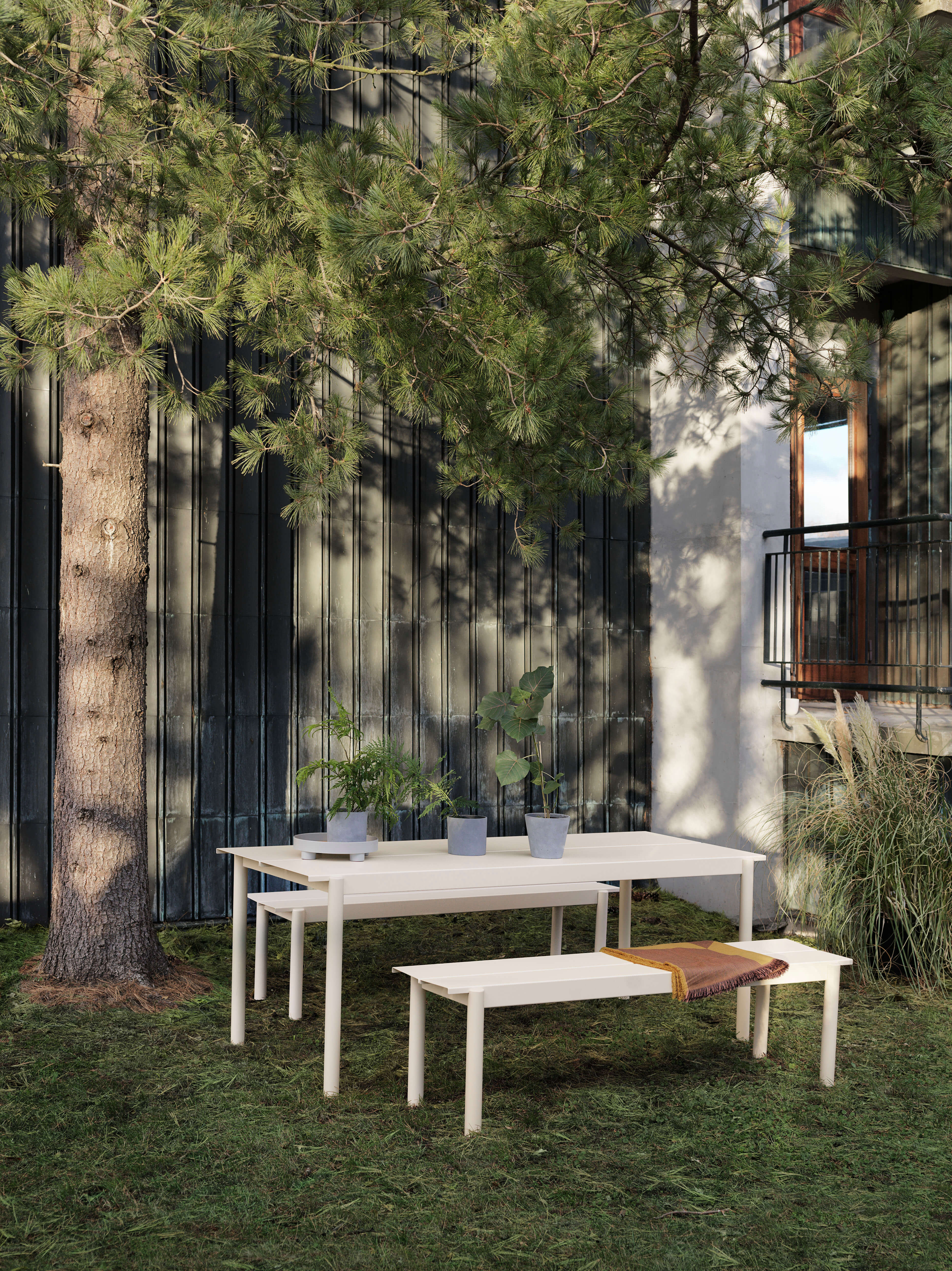 Outdoor Essplatz im Garten mit weissem Tisch und passenden Bänken auf Rasen. Auf dem Tisch stehen mehrere Topfpflanzen, im Hintergrund eine dunkle Holzfassade und ein Balkon neben einem grossen Baum.