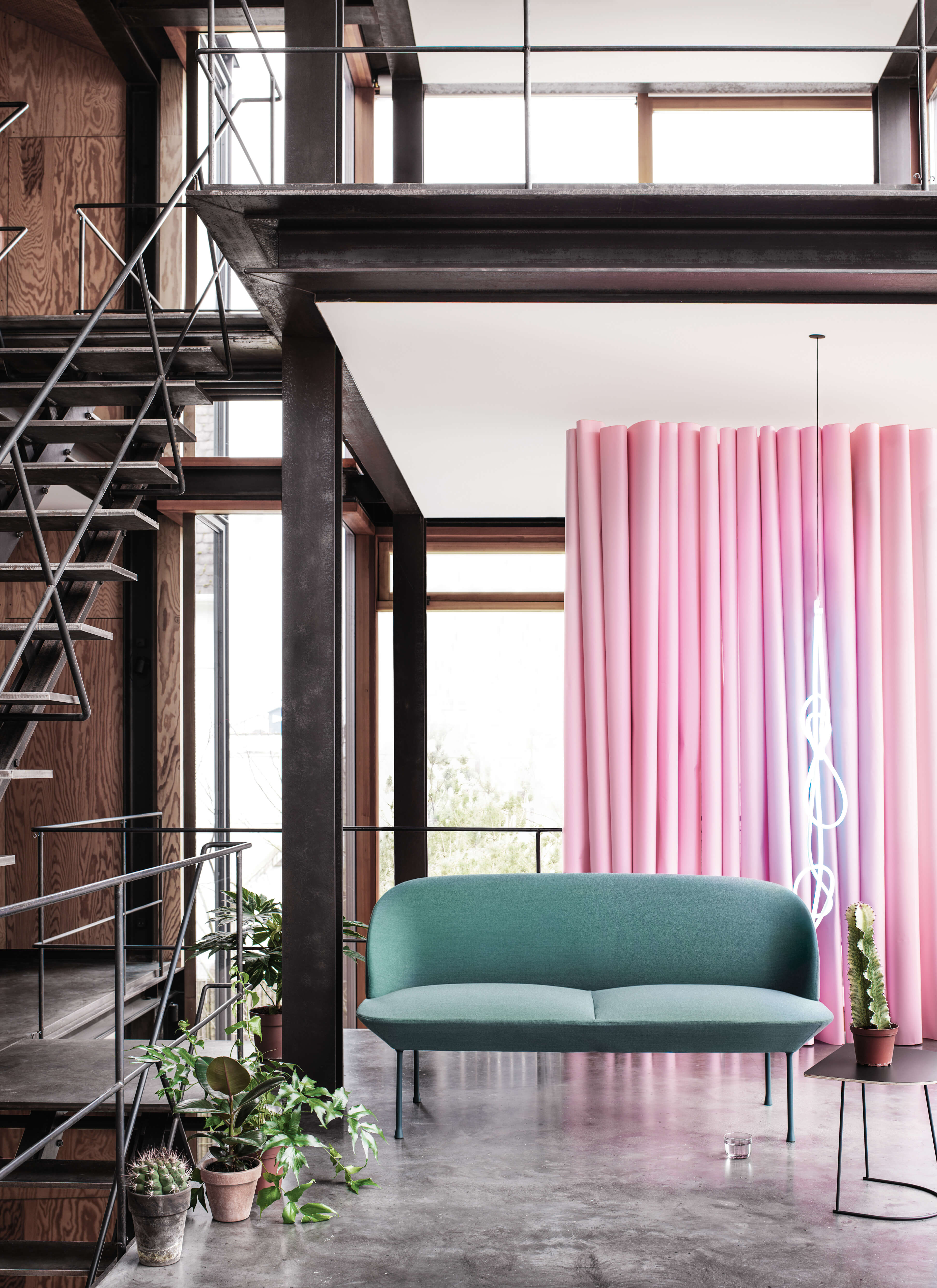 Industrial Loft Setting mit schwarzer Stahlkonstruktion und Treppe, Betonboden und grossen Fensterflächen. Im Fokus ein grünes Sofa vor einem rosa Vorhang, ergänzt durch mehrere Topfpflanzen.