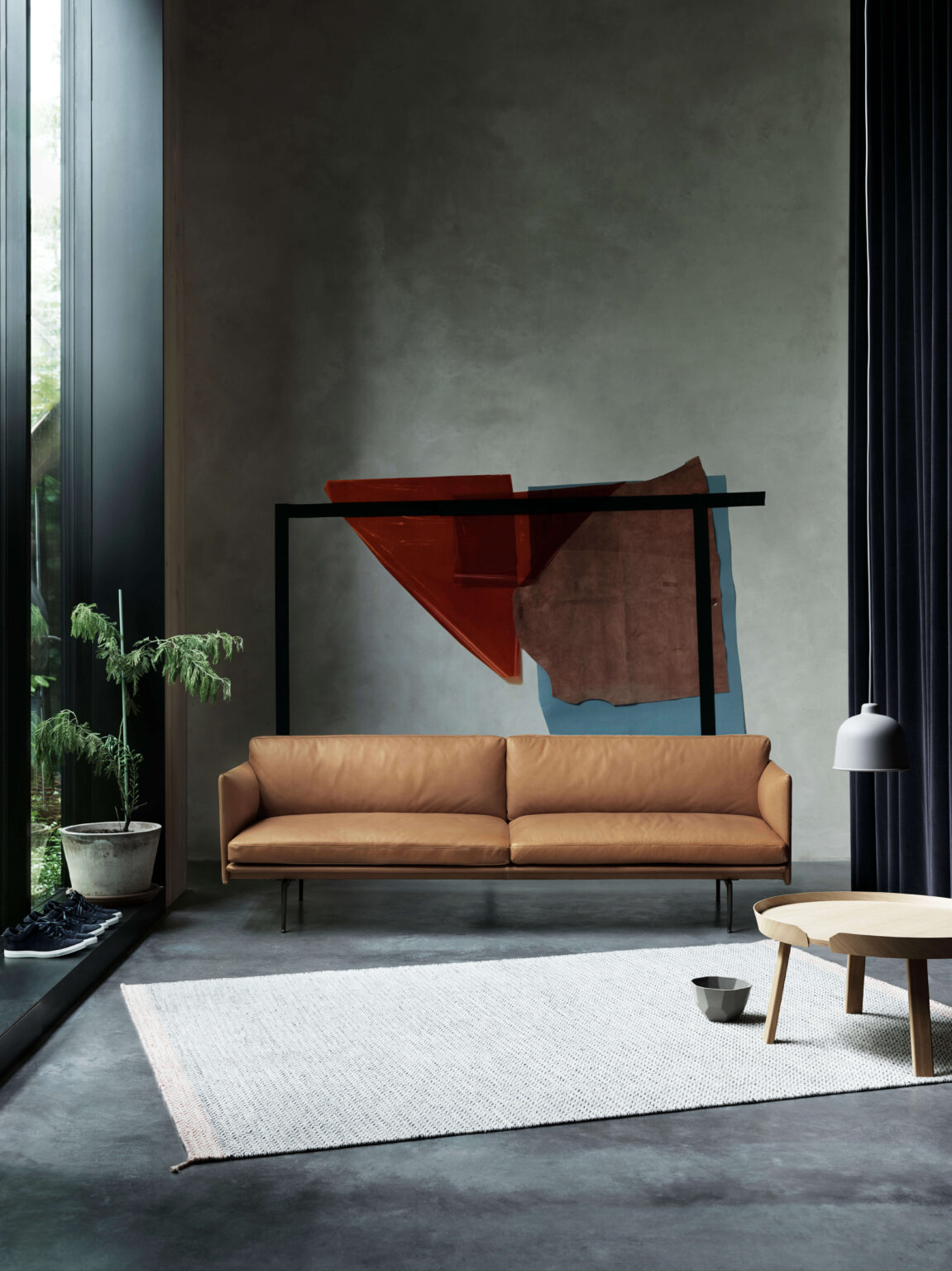 Modernes Wohnzimmer mit braunem Ledersofa vor grauer Wand, abstrakter Wandkunst, grossem Fenster, Pflanze, hellem Teppich und runden Beistelltischen.