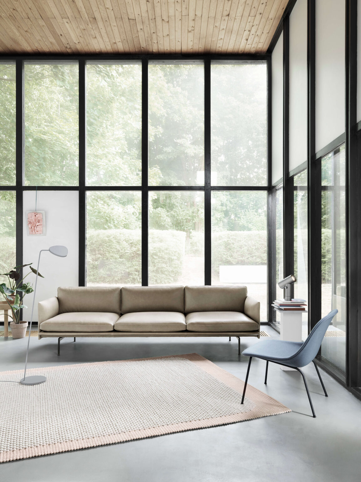 Helles Wohnzimmer mit bodentiefen Fenstern in schwarzen Rahmen, beigem Sofa auf hellem Teppich, blauem Stuhl, Stehleuchte und Pflanzen.